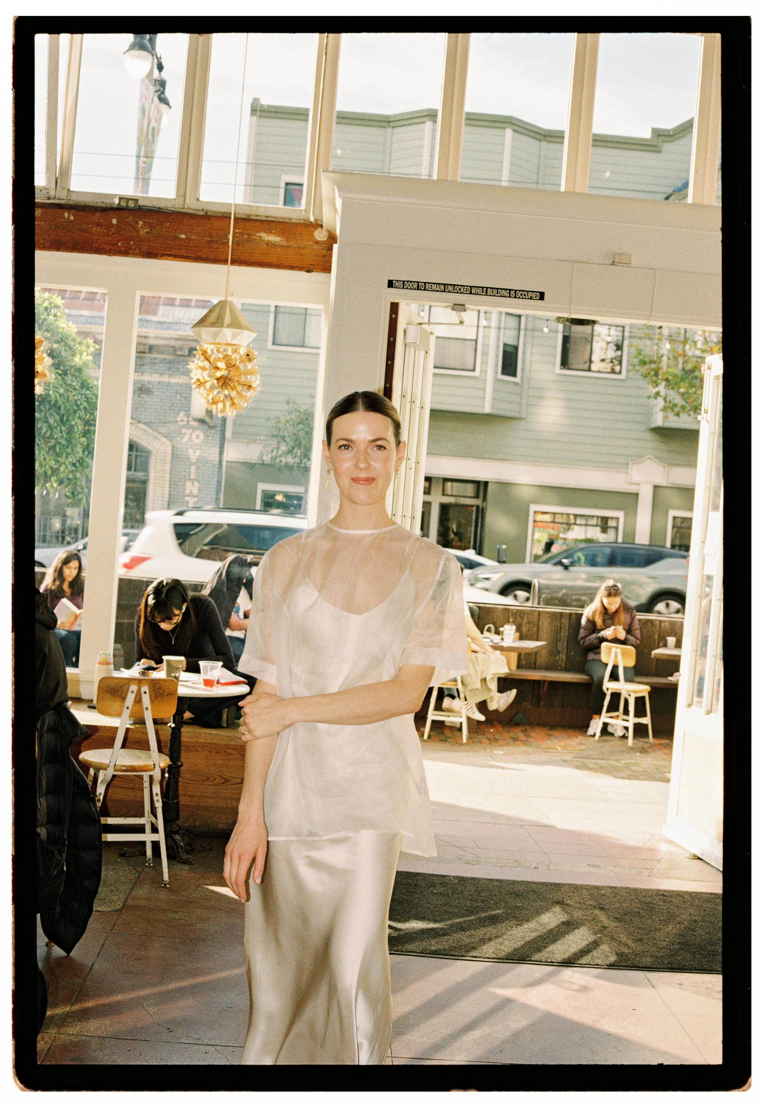 A woman standing inside a cafe or restaurant near the entrance, wearing a cream-colored silk dress with a sheer overlay, smiling at the camera. Through the open door and large windows, parked cars and people sitting outside are visible.
