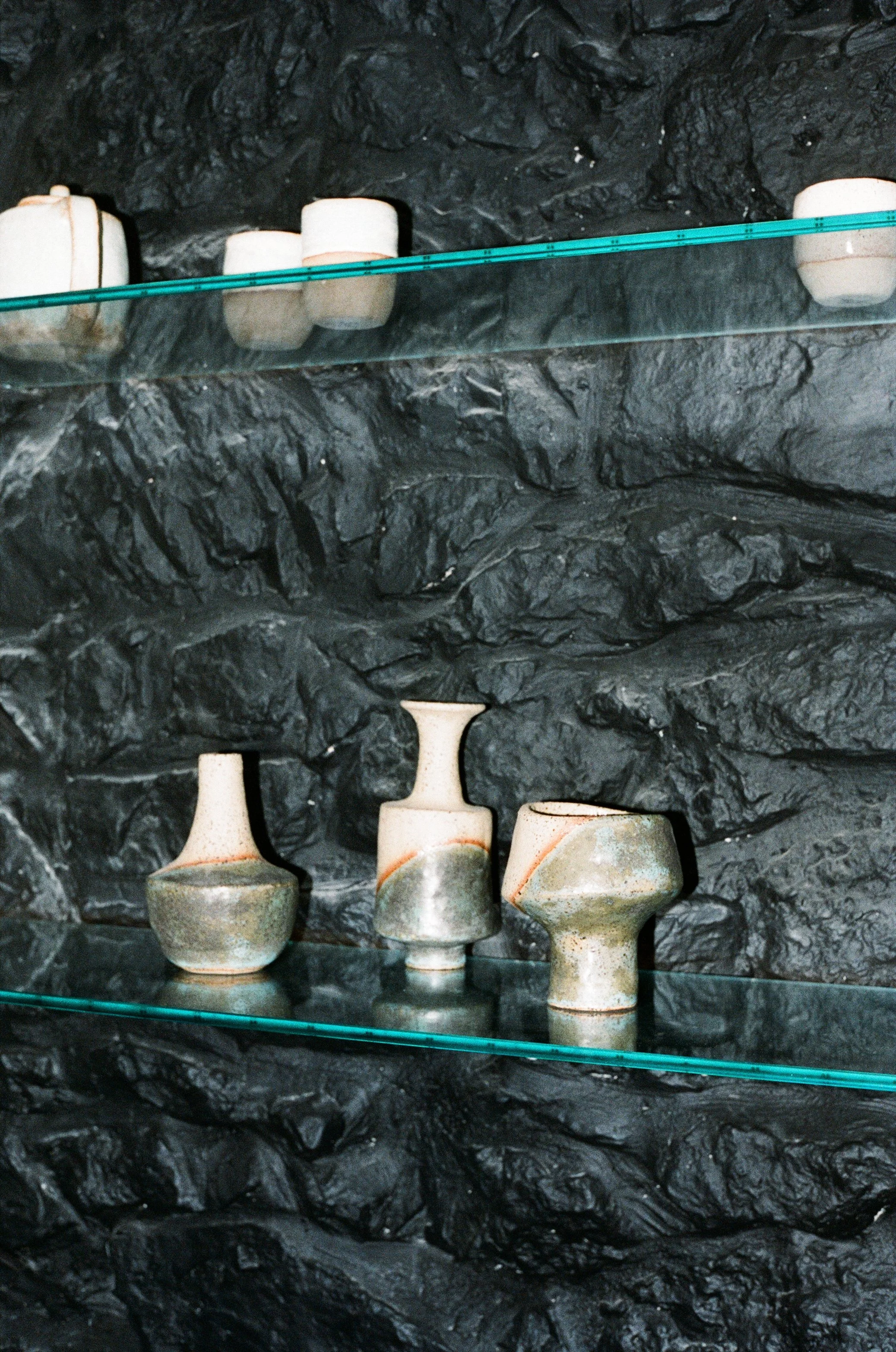 Artistic pottery displayed on glass shelves against a black textured rock wall.