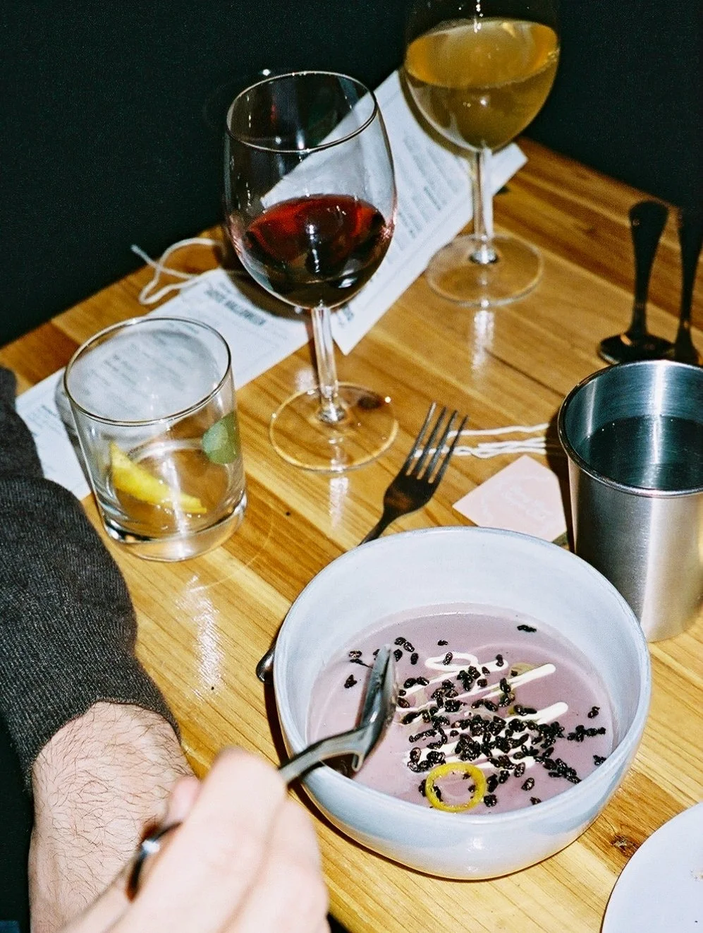 A table with a bowl of pink dessert topped with black sprinkles and a yellow ring, a person's hand holding a spoon, three glasses of wine (red, white, and yellow), a metal cup, a fork, a piece of paper, and a black object.