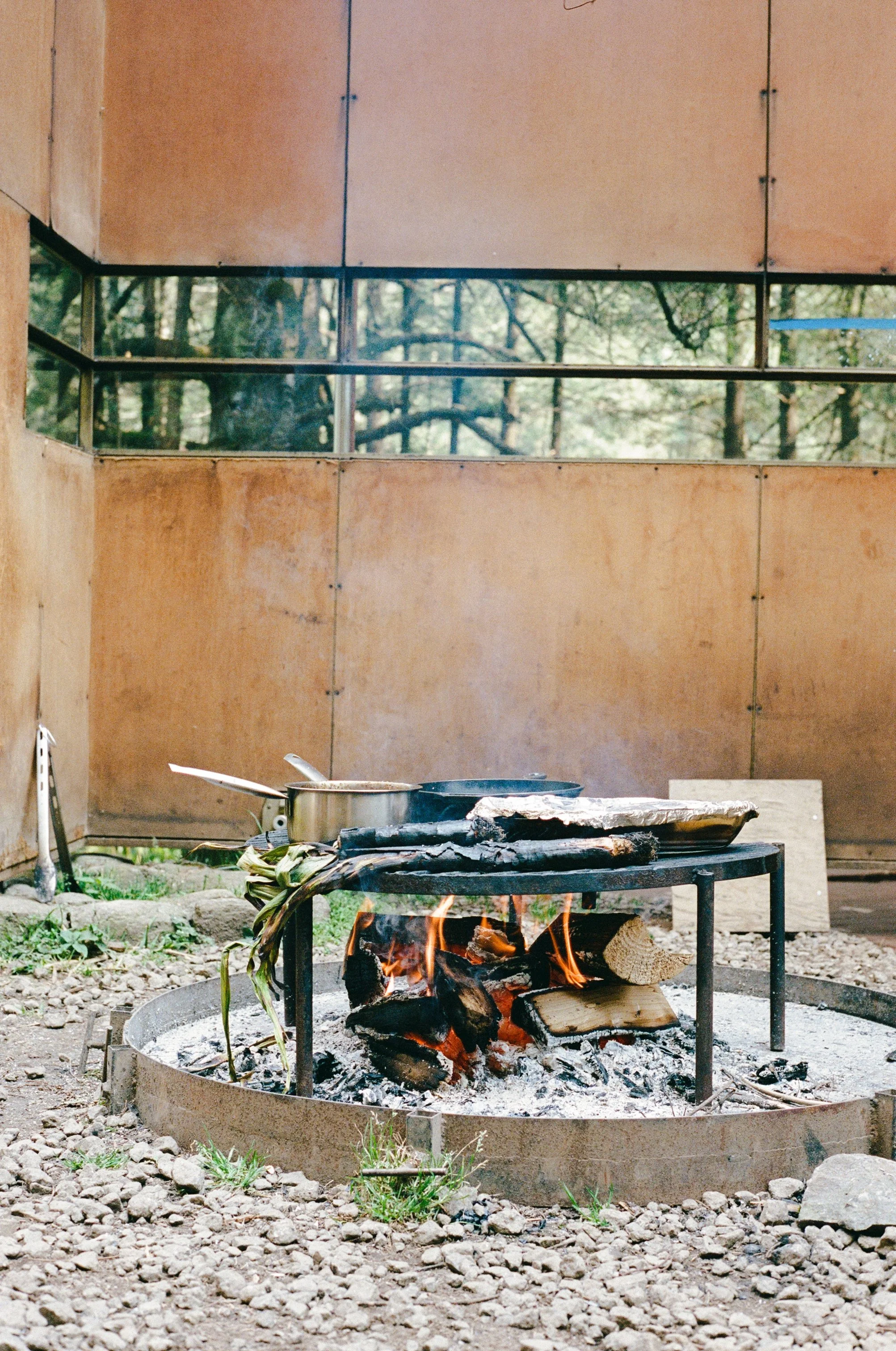 Outdoor fire pit with logs burning, cooking utensils, and a frying pan on a rustic table. Surrounded by a rocky ground with a wooden wall and trees in the background.