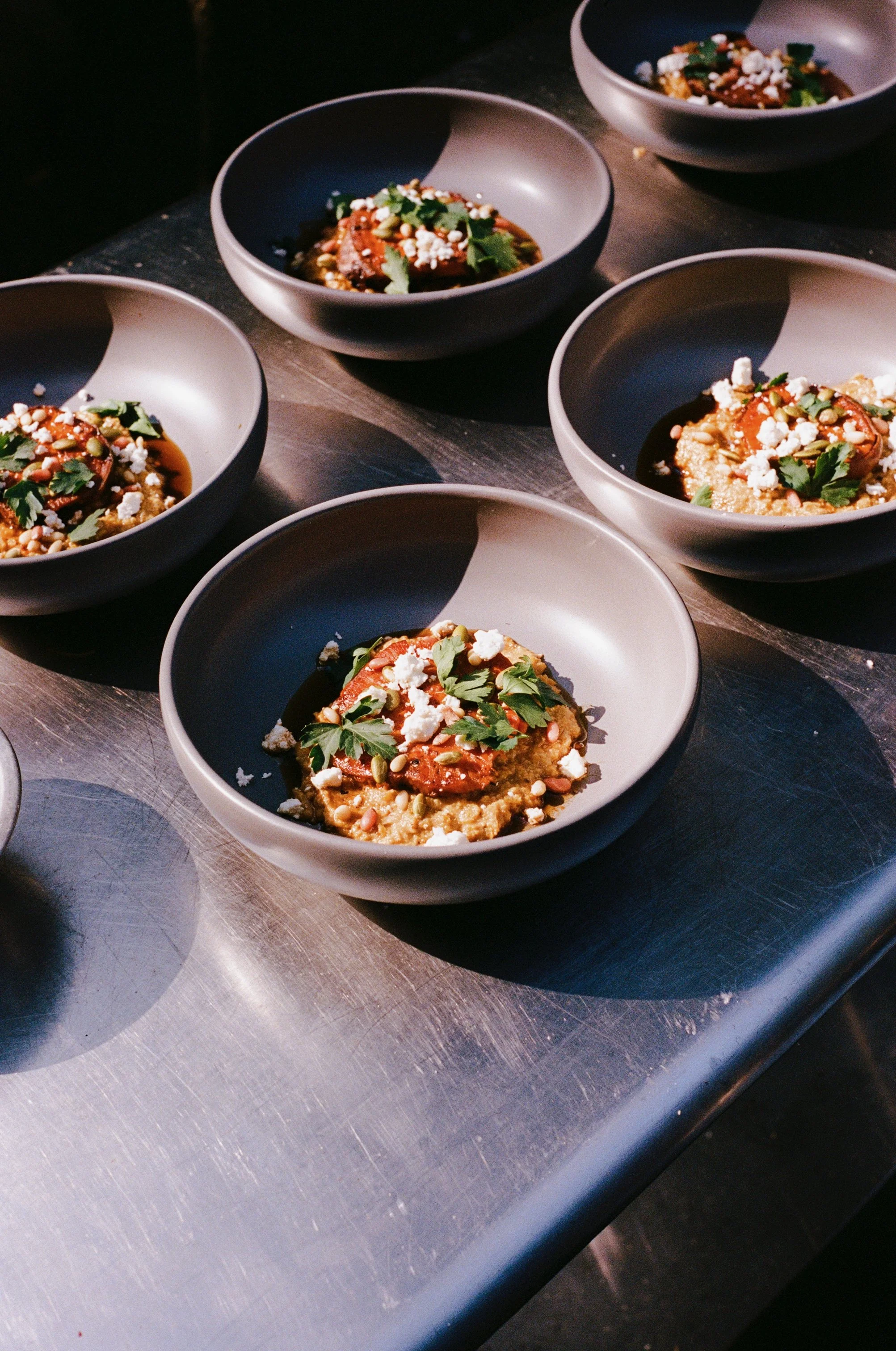 Six bowls of a young cornmeal dish garnished with cilantro, cotija cheese, and seeds on a metal countertop.