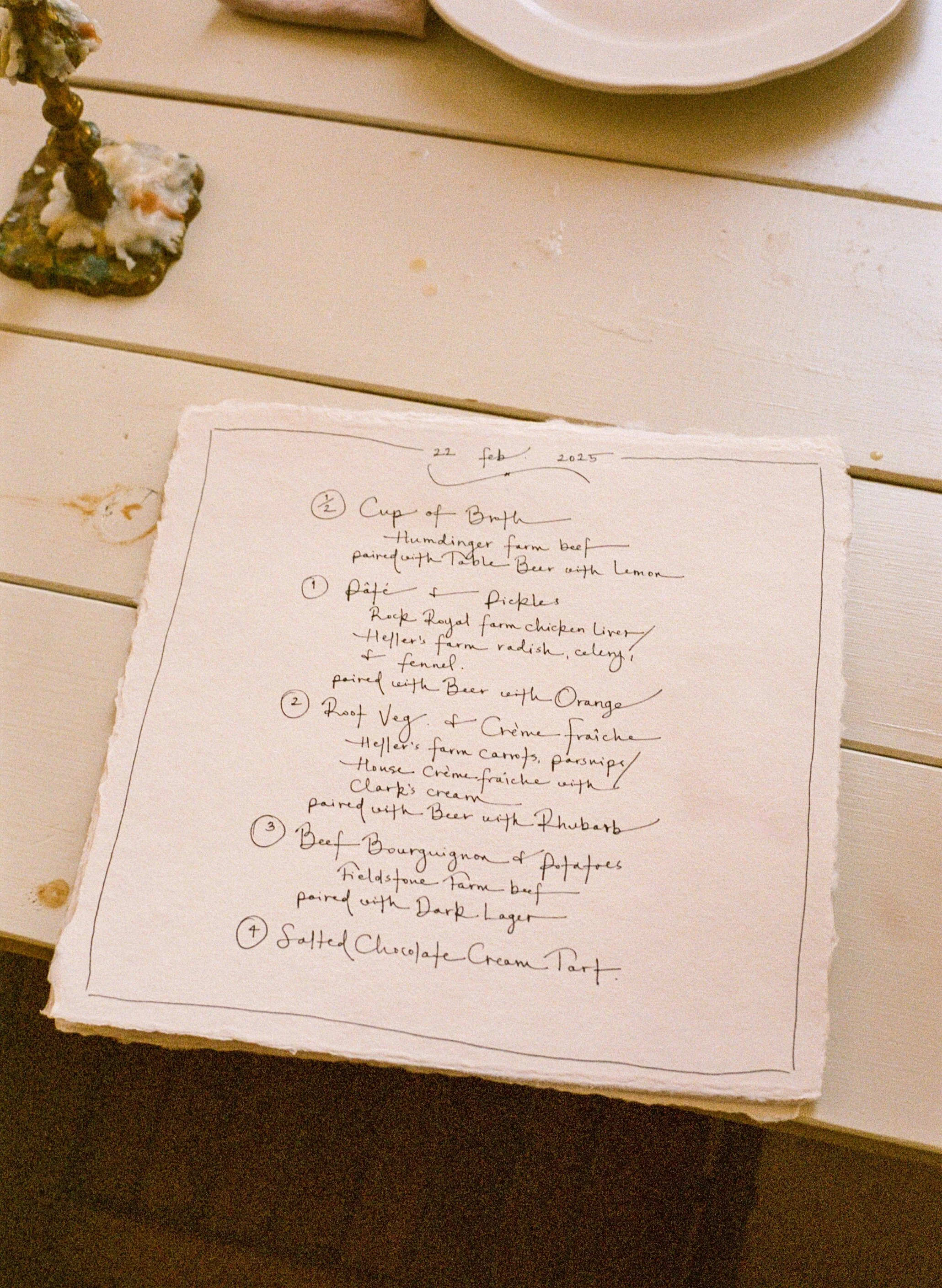 Handwritten recipe or menu on a torn piece of paper, listing dishes with numbers, set on a white wooden table with a blank plate nearby.