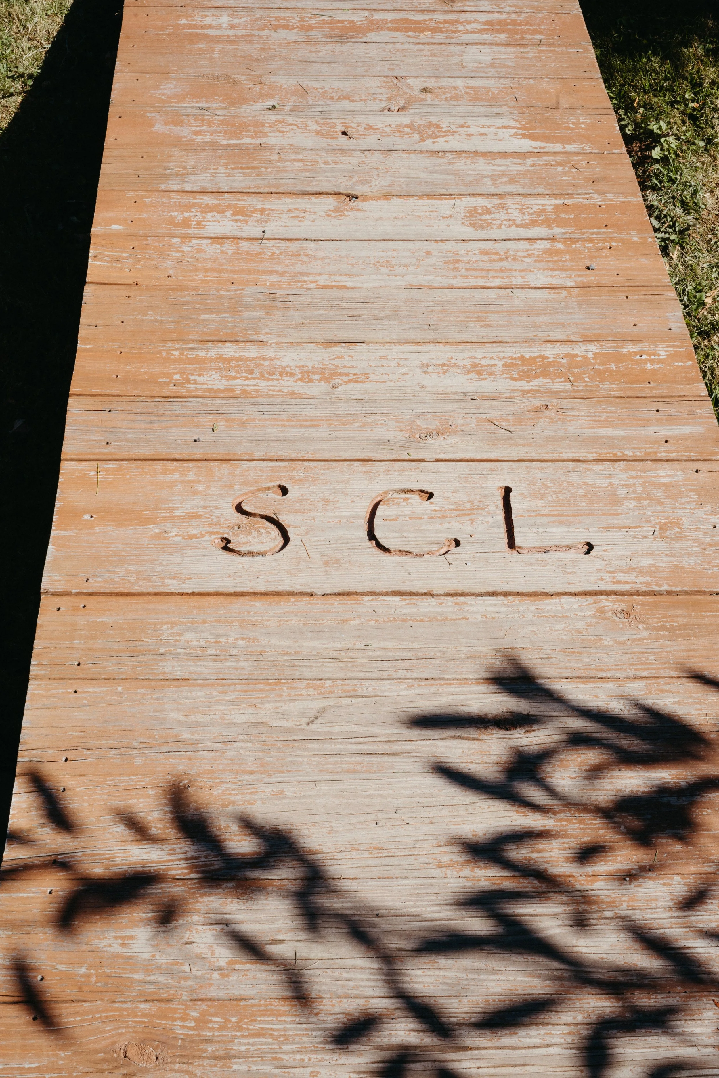 Wooden board with the initials 'S C L' carved into it, outdoor setting with shadow of a tree branch on the lower part.