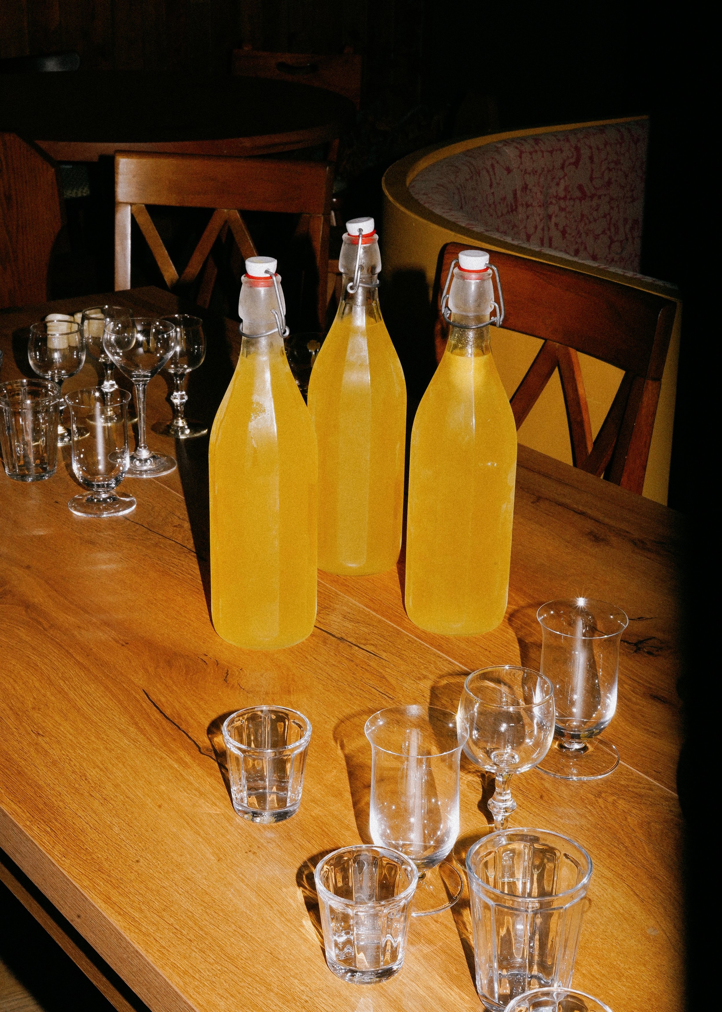 Three glass bottles filled with limoncello are on a wooden table, surrounded by various empty drinking glasses and shot glasses. The setting is indoors in a restaurant or bar.