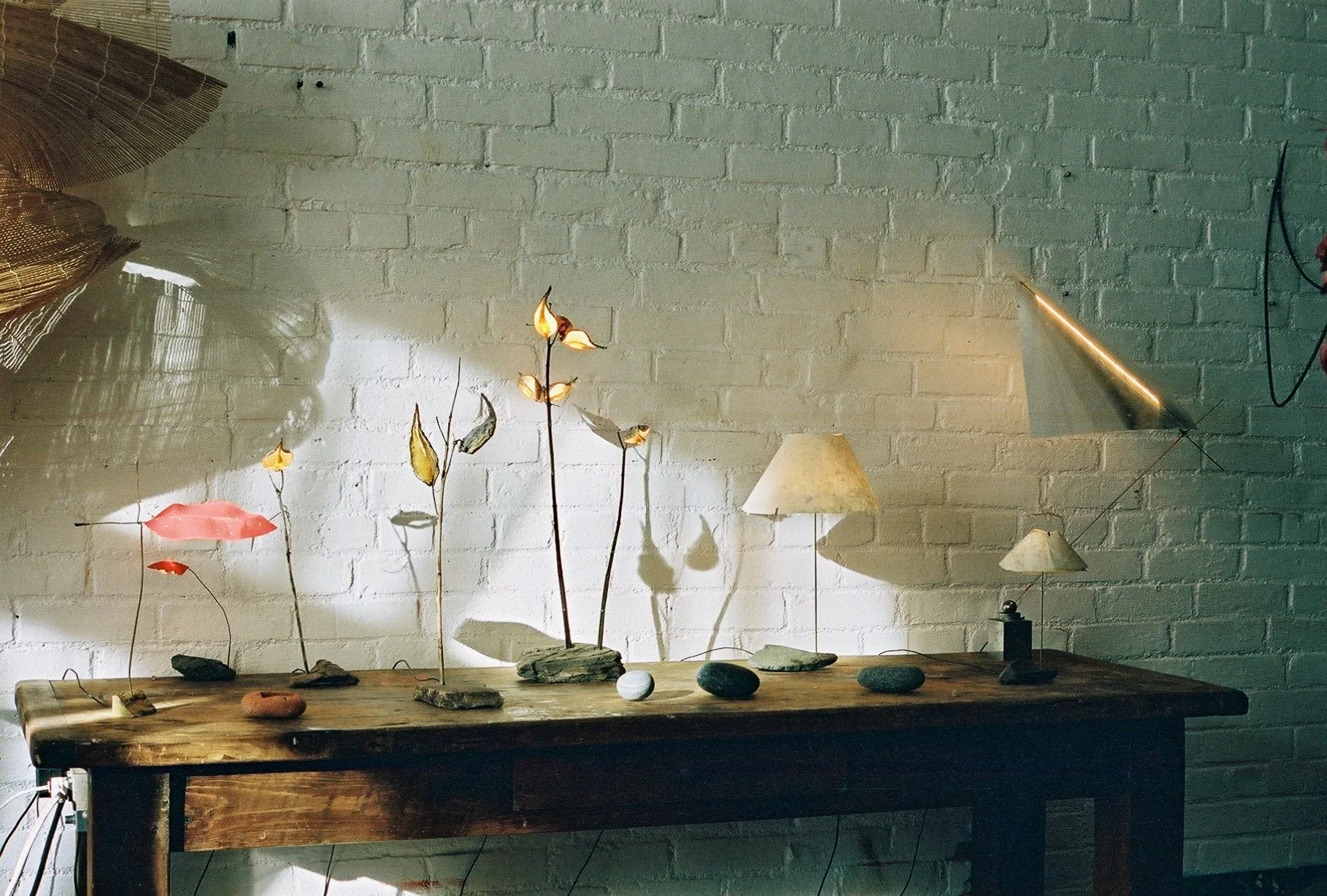 Decorative table with ceramic flower sculptures, lamps, and stones against a white brick wall with shadows and lighting.