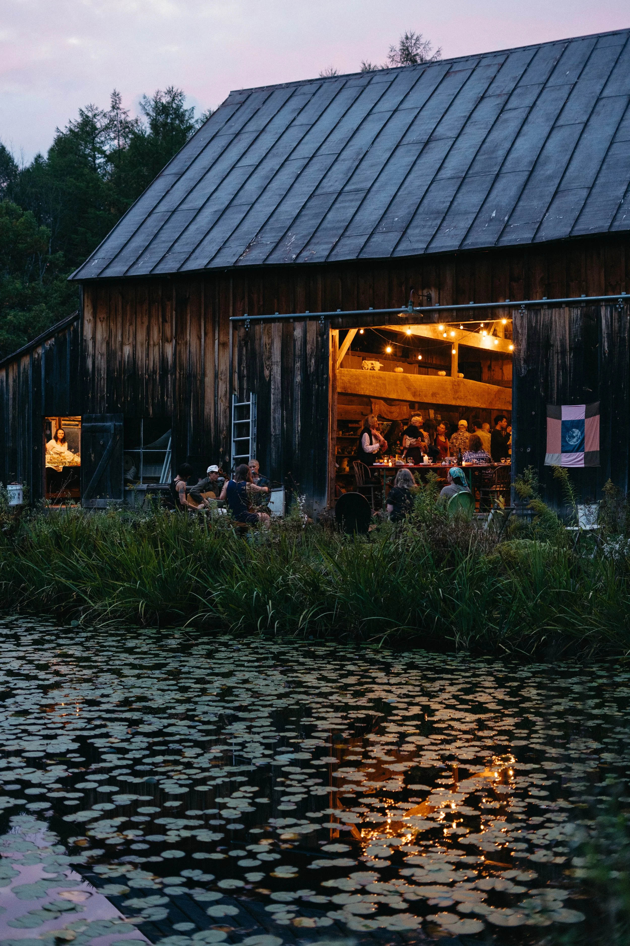 People gathered inside a barn decorated with string lights for an evening event, overlooking a pond with water lilies.