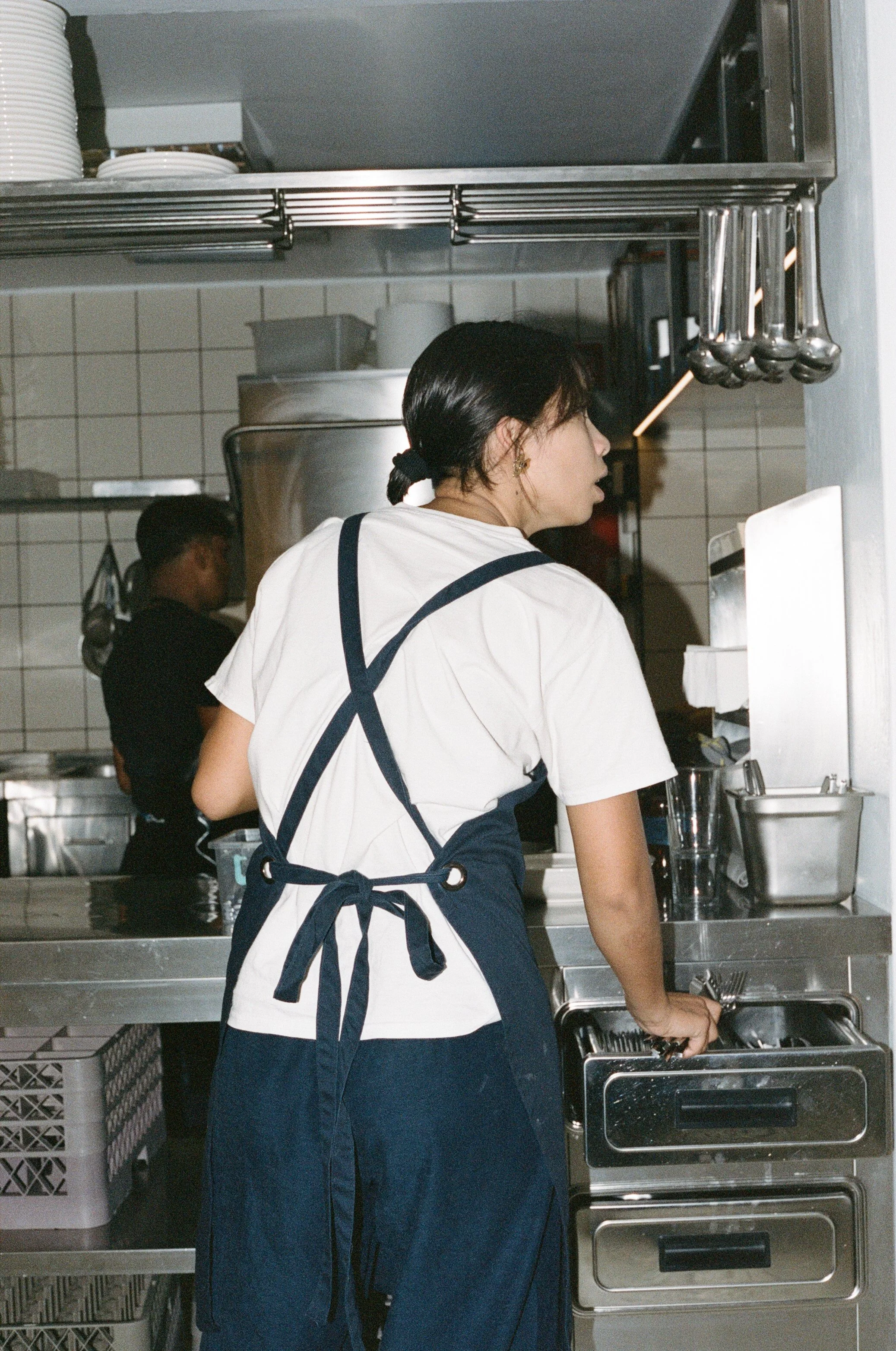 A woman in a white t-shirt and navy apron, with black hair tied in a ponytail, working in a commercial kitchen.