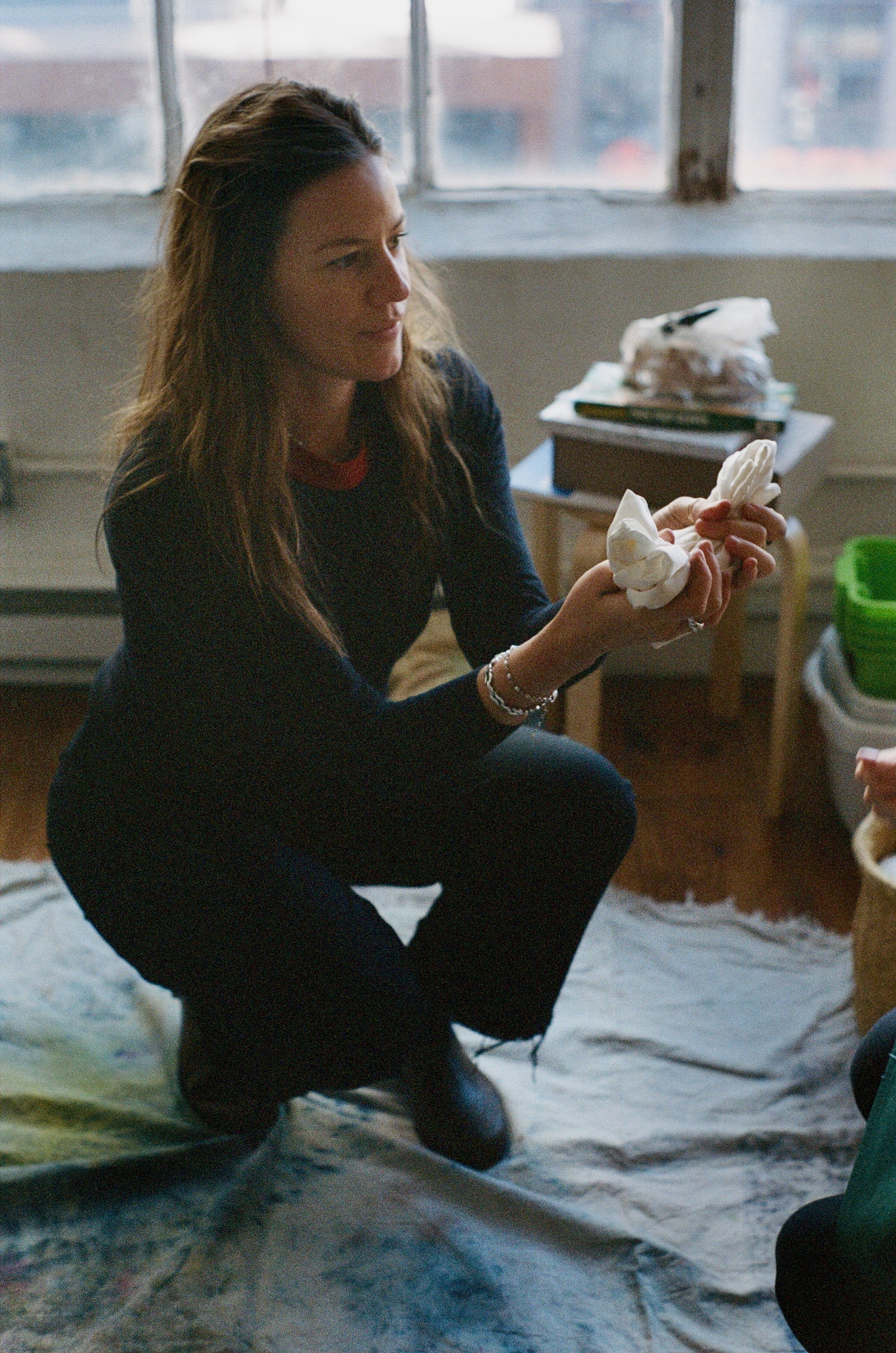A woman with long brown hair in black clothing squats on the floor inside, holding a cloth in her hands.