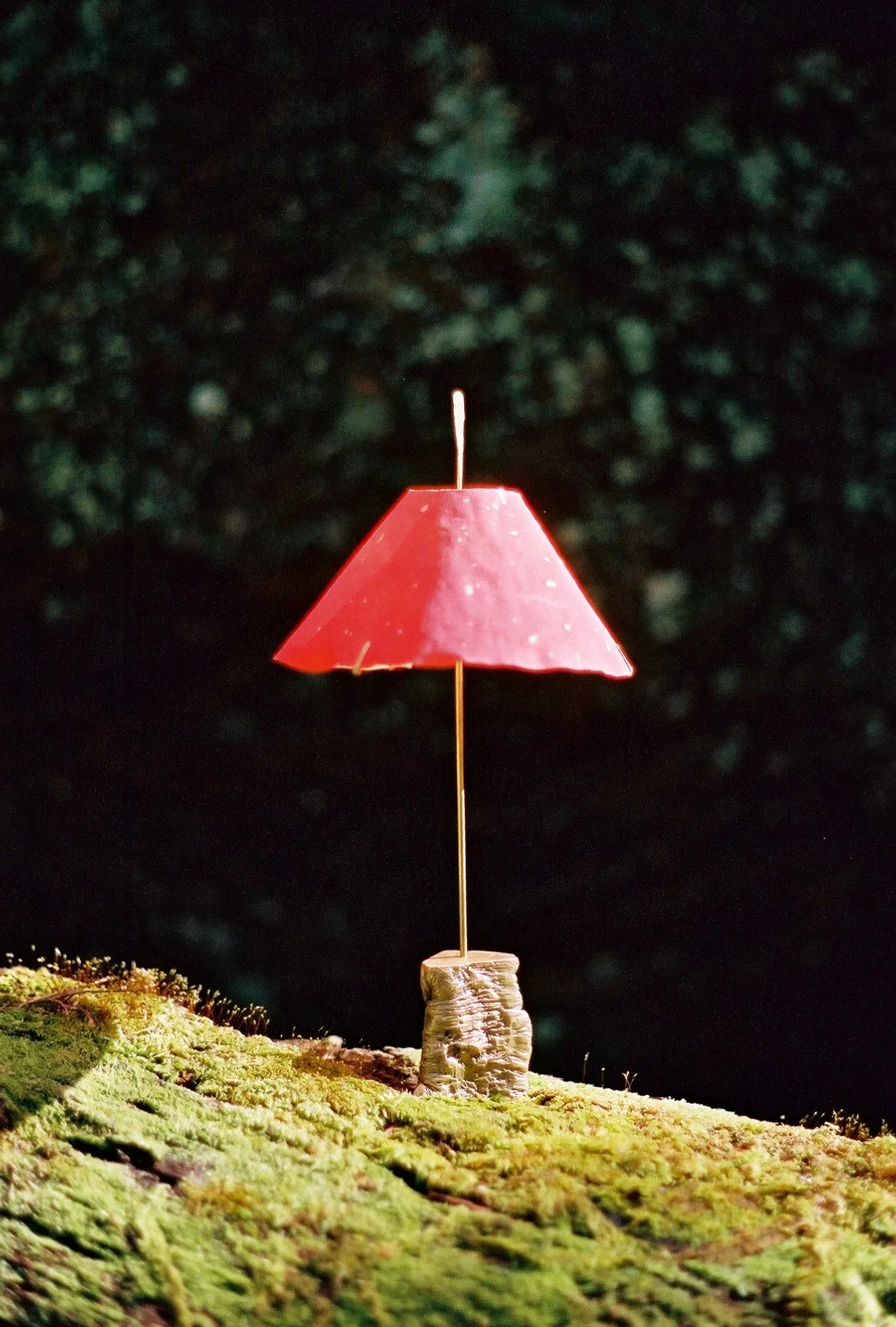 A miniature umbrella with a red canopy and a wooden handle is placed on a mossy surface, with a dark blurred background.
