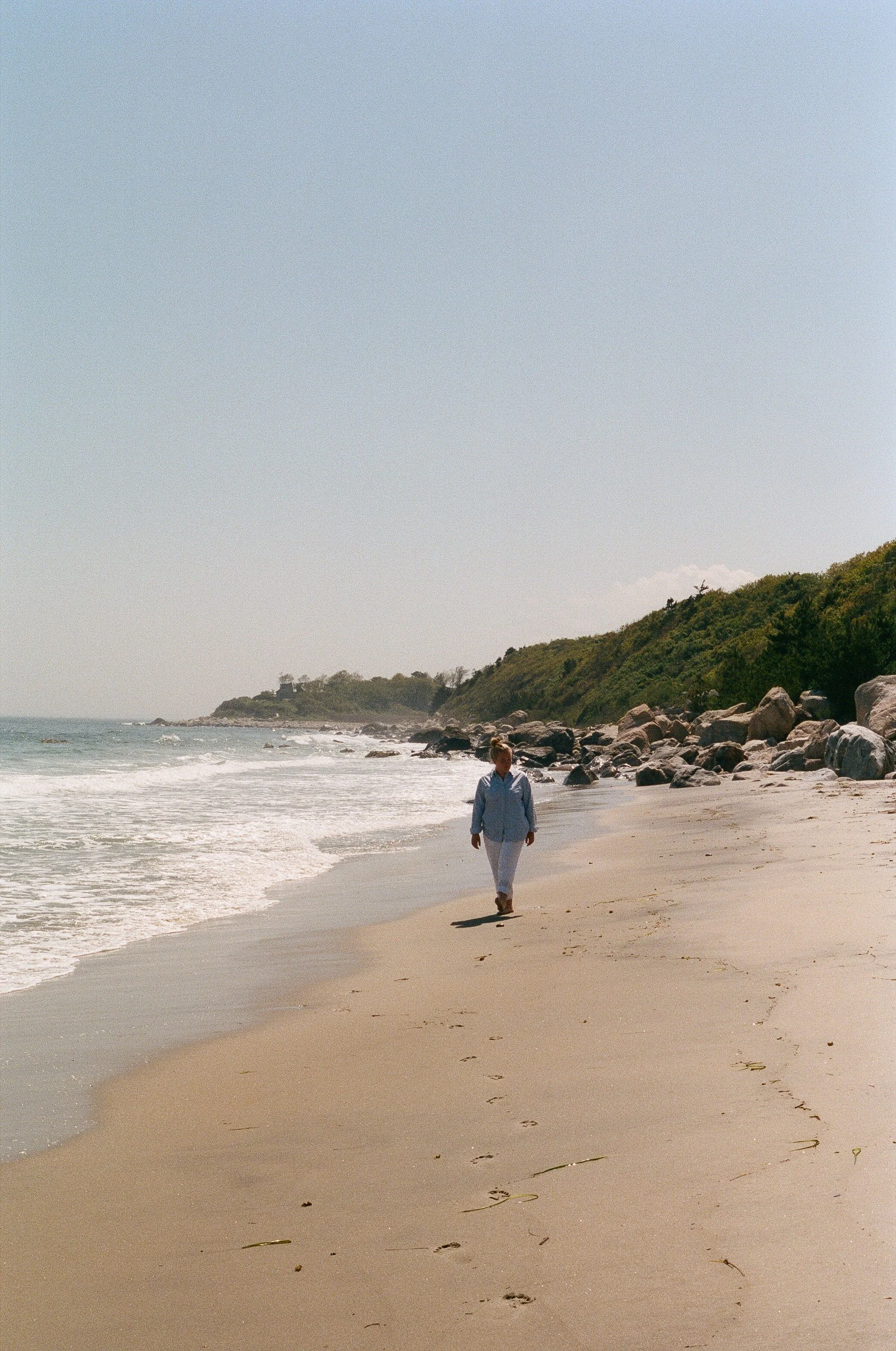 A woman walking alone along a sandy beach near the ocean, with green hills and rocks in the background on a clear day.