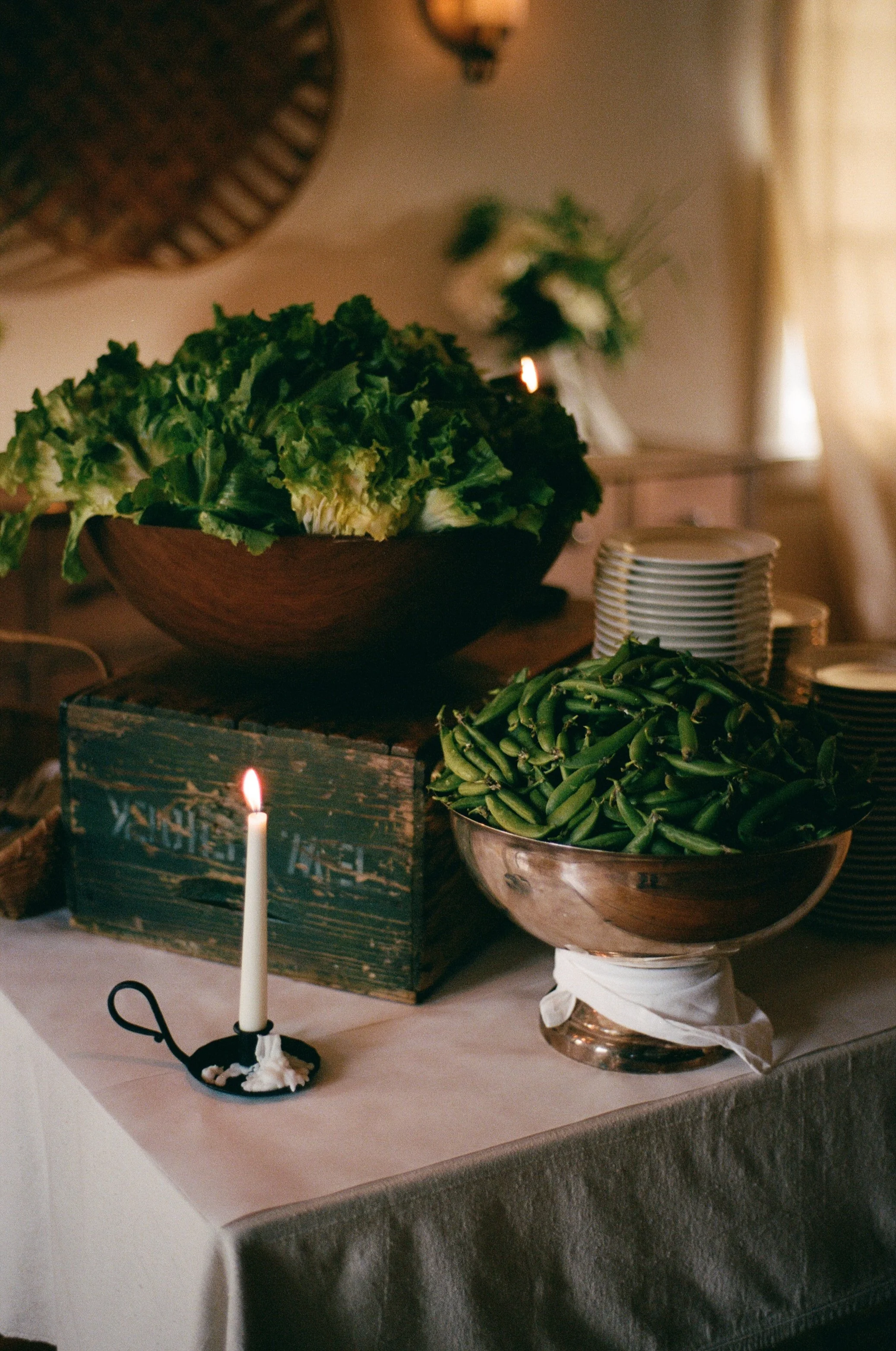 A table with bowls of green leafy lettuce and snap peas, lit candles, and stacked plates in a softly lit room.