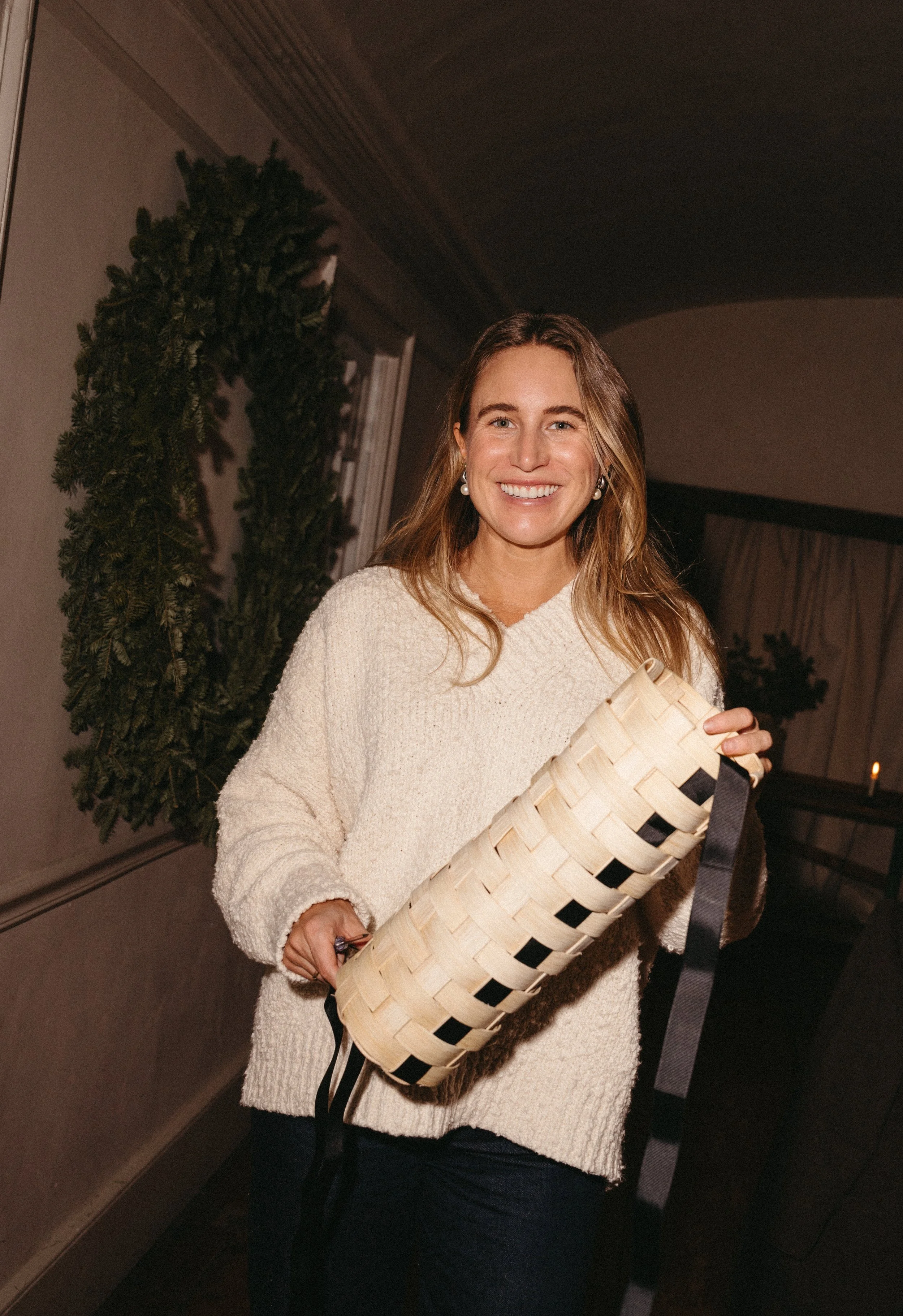 A woman with long brown hair, smiling and holding a woven basket, standing indoors with a Christmas wreath and curtains in the background.