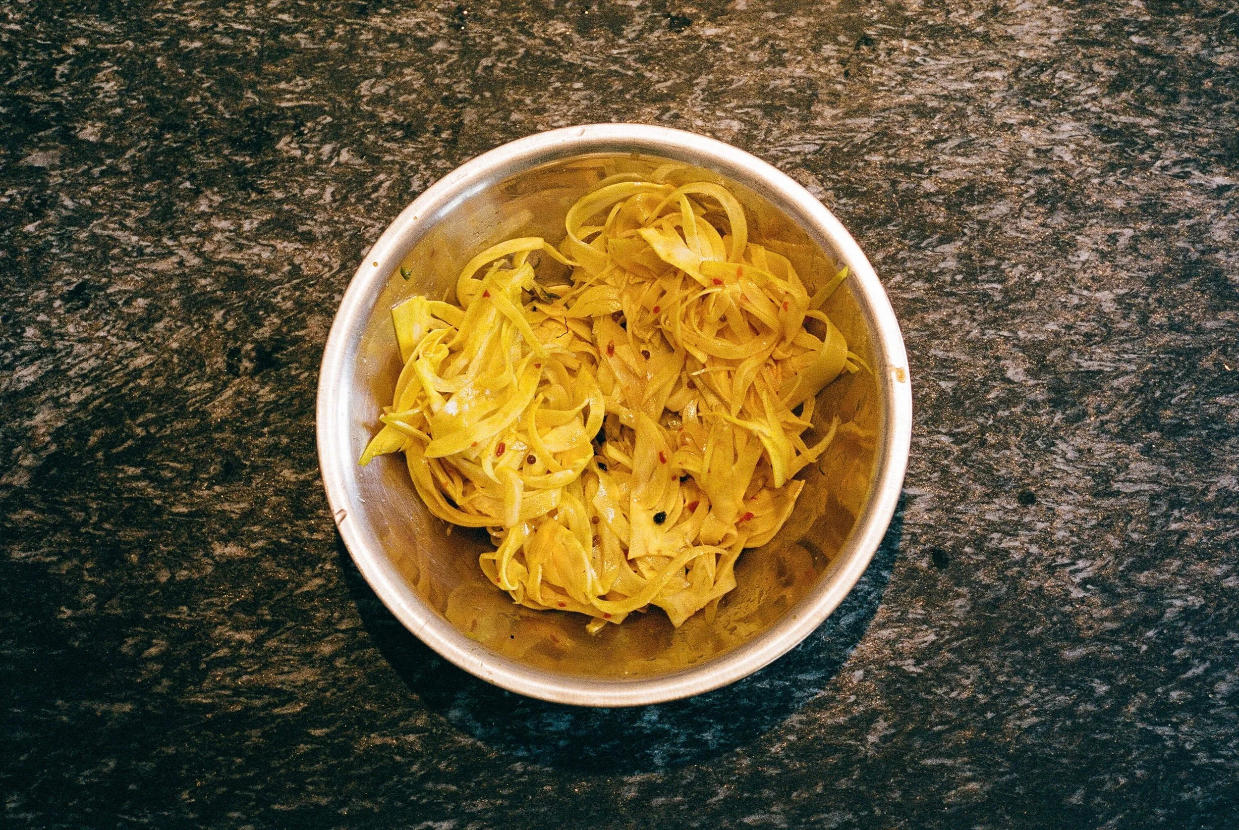 A metal bowl containing yellow pickled vegetables on a black, textured countertop.