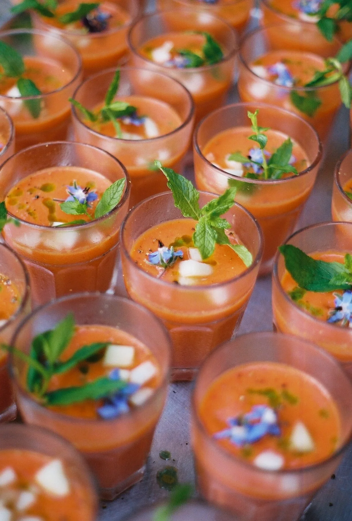 Multiple small glasses filled with orange-colored soup or beverage, garnished with green herbs, edible flowers, and small white pieces, arranged on a surface.