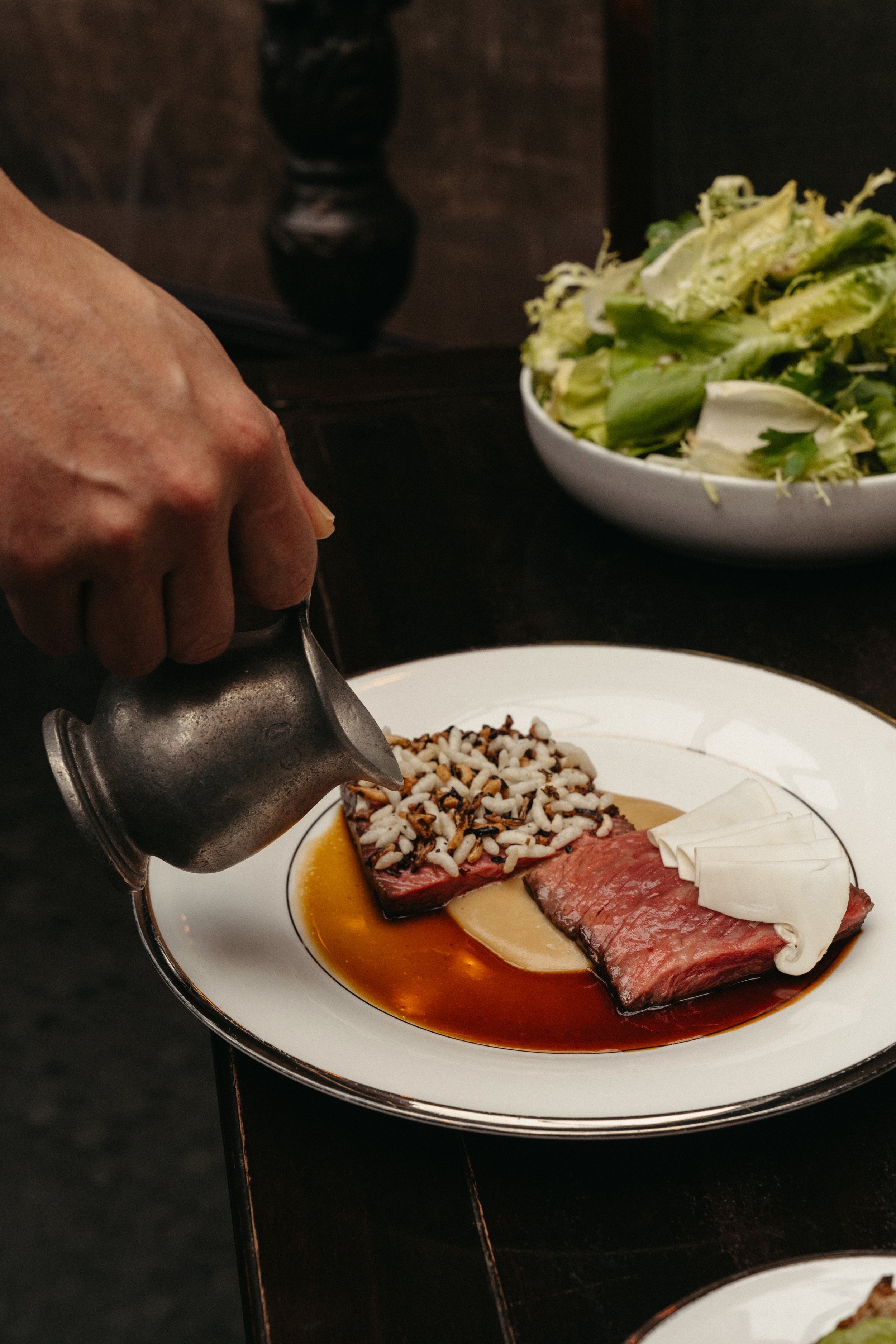 Close-up of a person pouring sauce over seared beef topped with crunchy garnishes, served with sauce, on a white plate; a bowl of salad in the background.