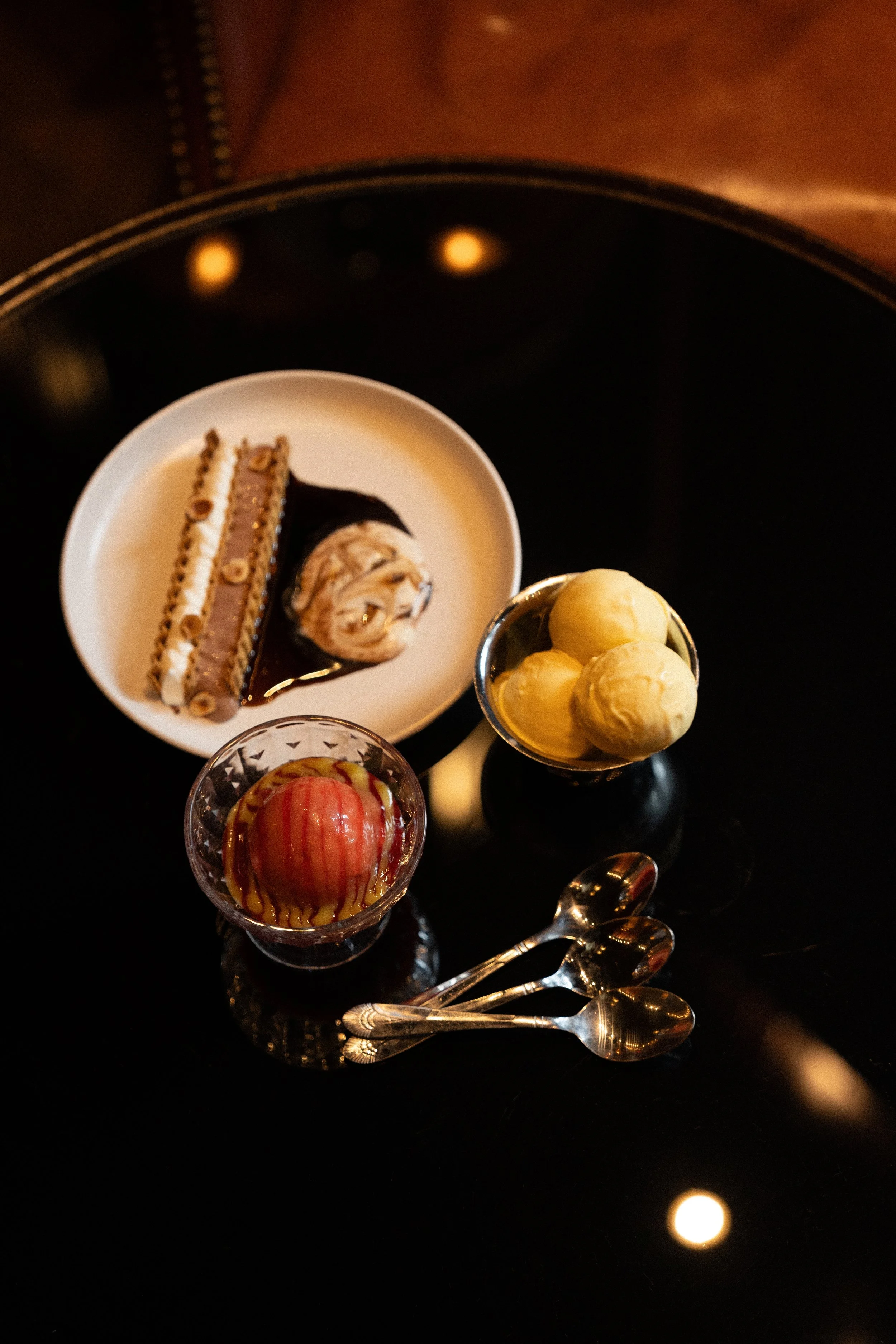 A selection of three small desserts on a black reflective surface with three small spoons. The desserts include a red apple-shaped treat with caramel drizzle, three scoops of yellow ice cream, and a white plate with a layered chocolate and cream cake