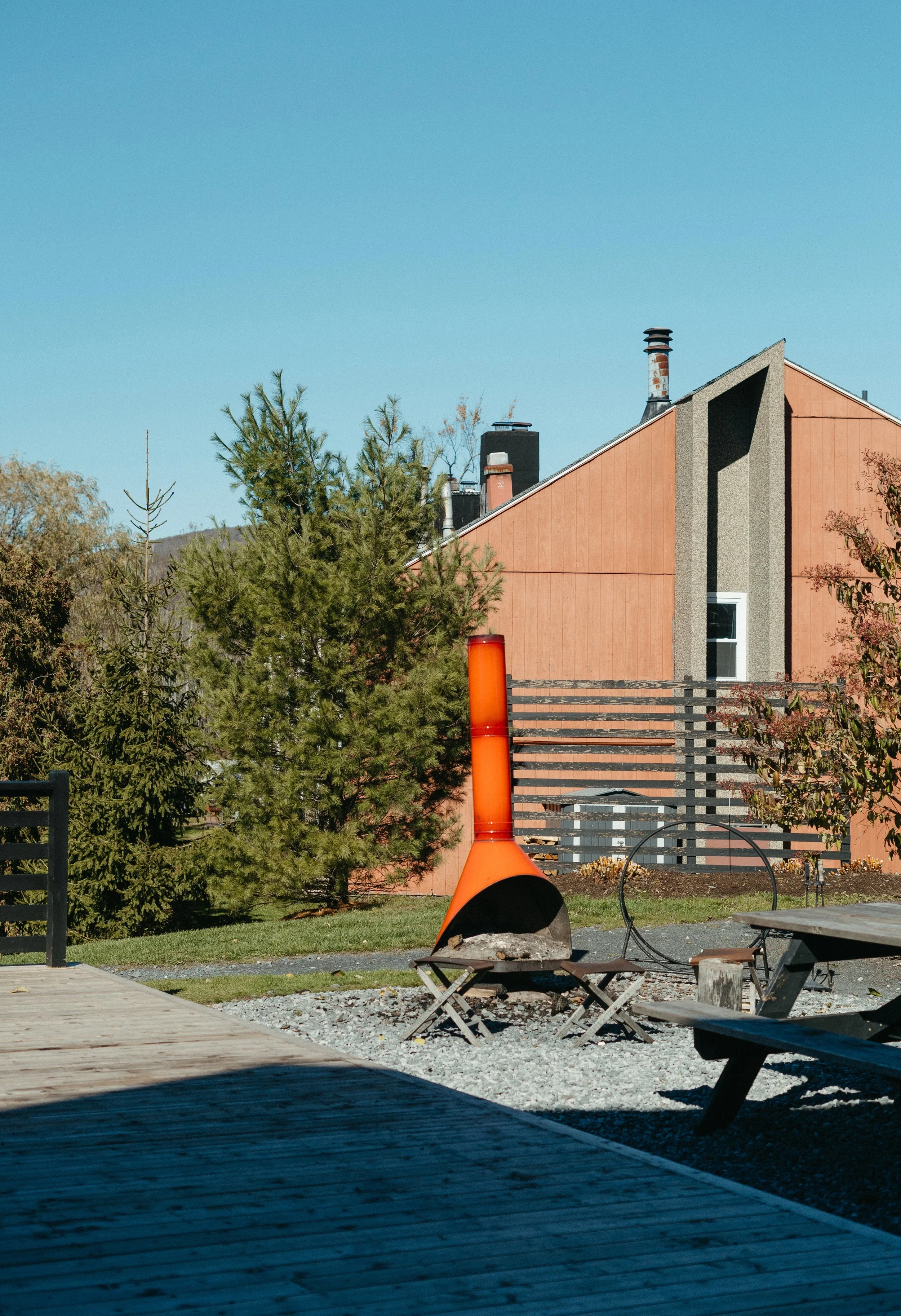 An outdoor patio area with a wooden deck, picnic tables, and a vintage sculptural orange metal fireplace. There are trees and a modern-style house with a reddish facade and black chimneys in the background, under a clear blue sky.
