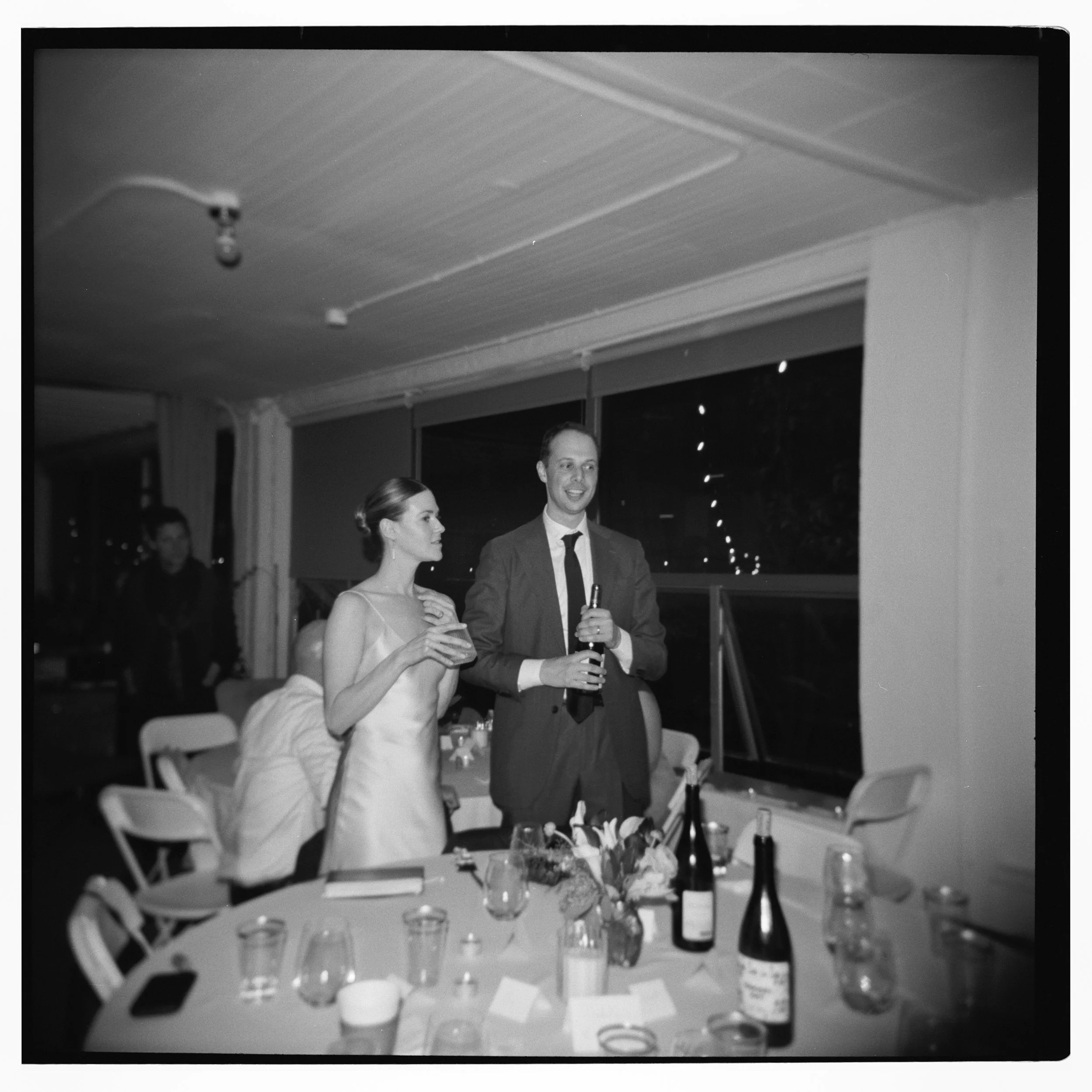 Black and white photo of a woman and man at a celebration dinner, with the man holding a bottle and smiling, and the woman standing beside him. The table nearby has wine bottles, glasses, and floral decorations.