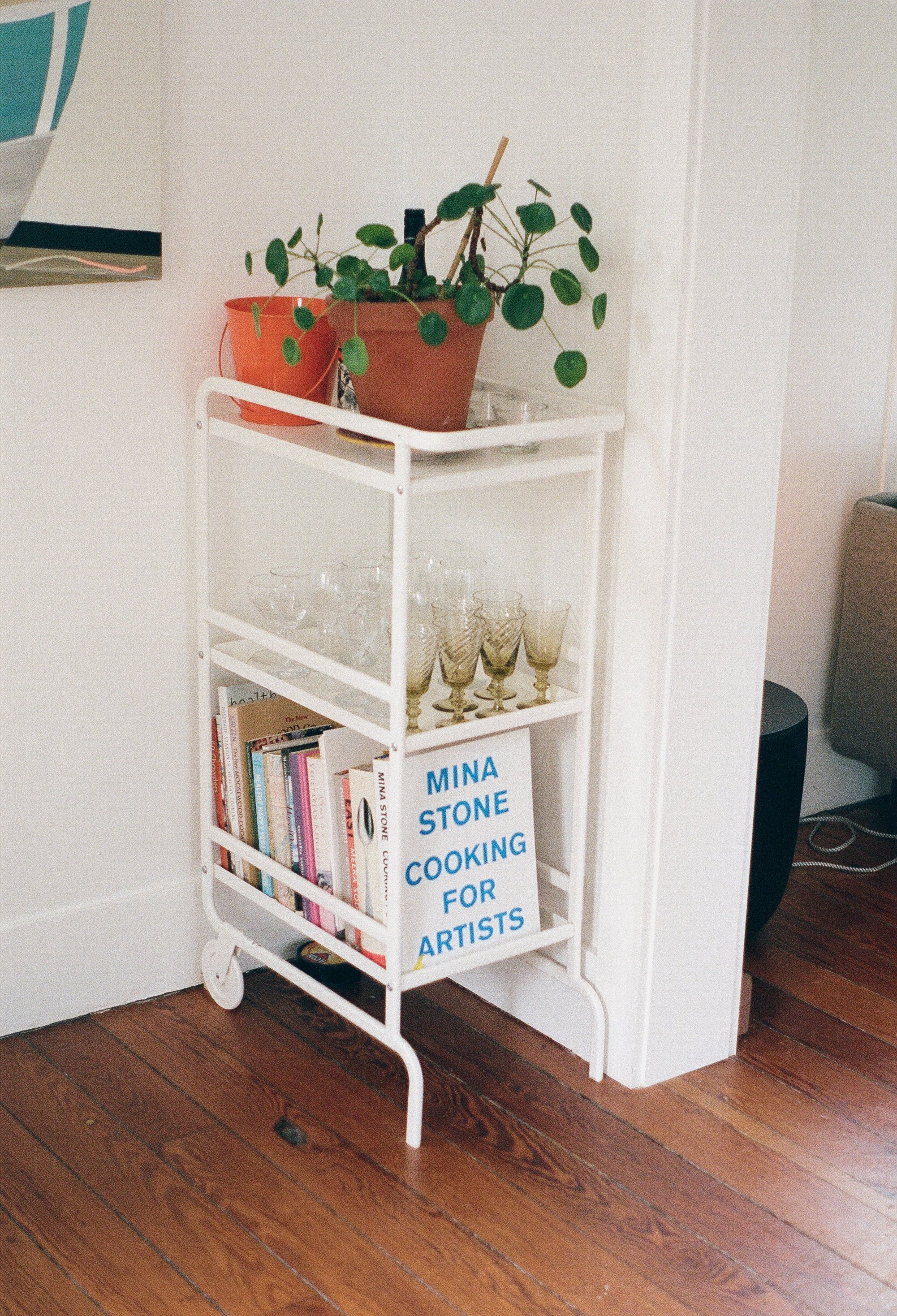 A white rolling cart with three shelves, holding books, glassware, and a sign that reads 'MINA STONE COOKING FOR ARTISTS', placed against a white wall on a wooden floor. The top shelf has potted plants and a small tray.