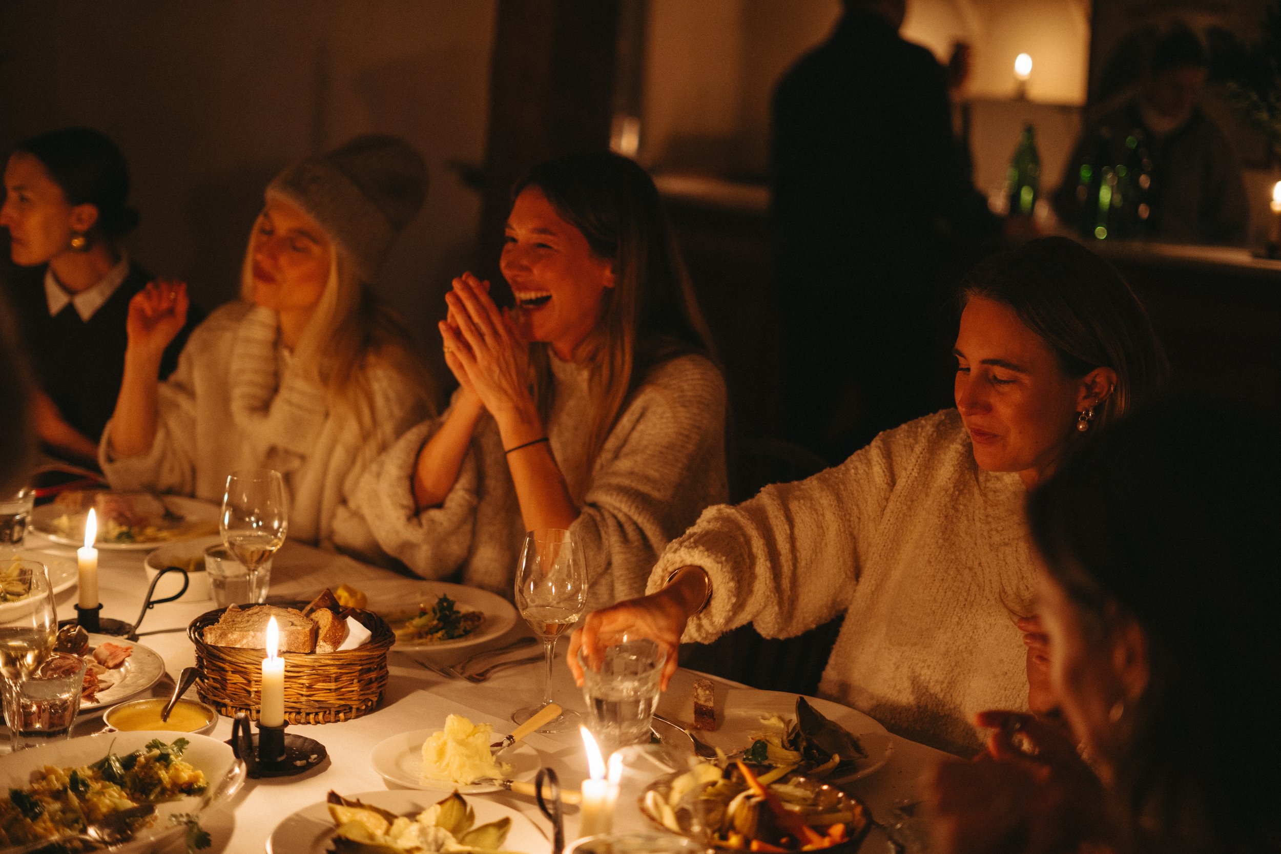 People enjoying a dinner party in a warm, dimly lit room with candles on the table, sharing smiles and laughter.
