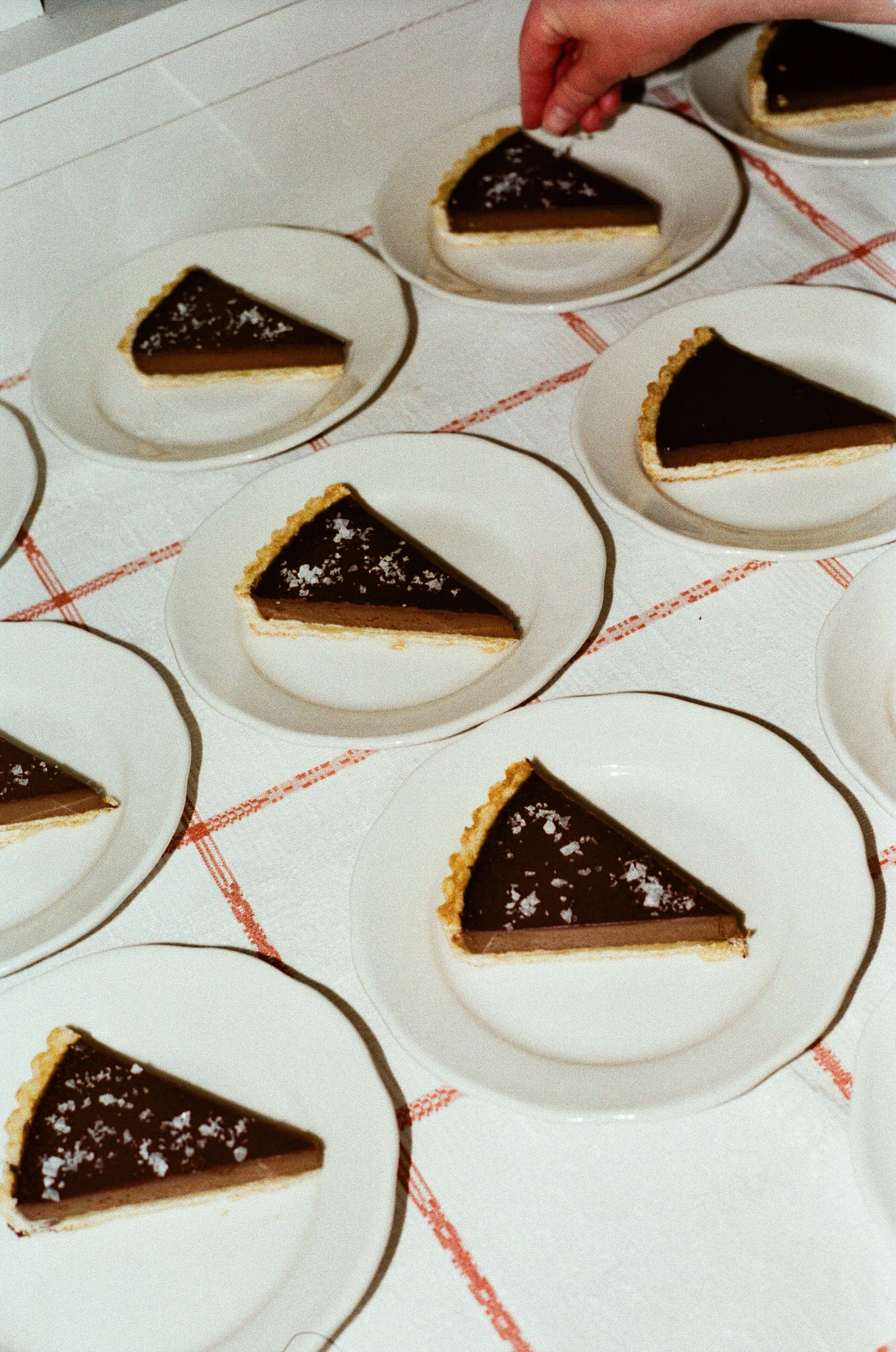 Multiple slices of chocolate pie on white plates arranged on a checkered tablecloth, with a hand placing one slice onto a plate.