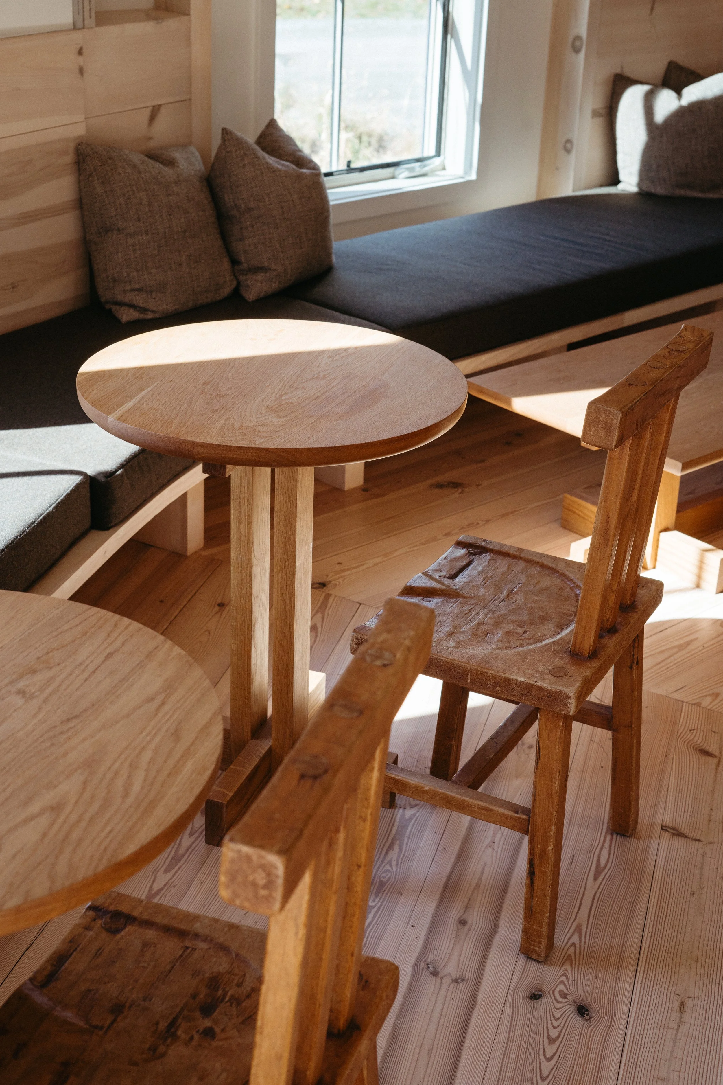 Interior of a wooden room with a window seat, small round wooden tables, and wooden chairs.
