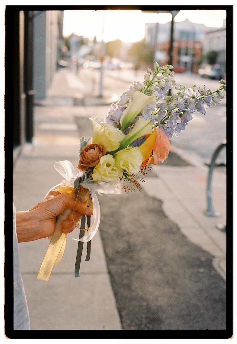 Person holding a small bouquet of mixed flowers on a city sidewalk during sunset.
