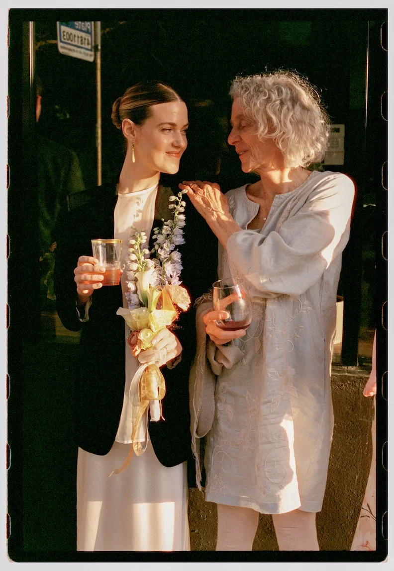 A young woman in a black blazer and white dress holding a floral bouquet and a glass of wine, standing next to an older woman with curly white hair in a light-colored dress, sharing a moment during an outdoor evening gathering.