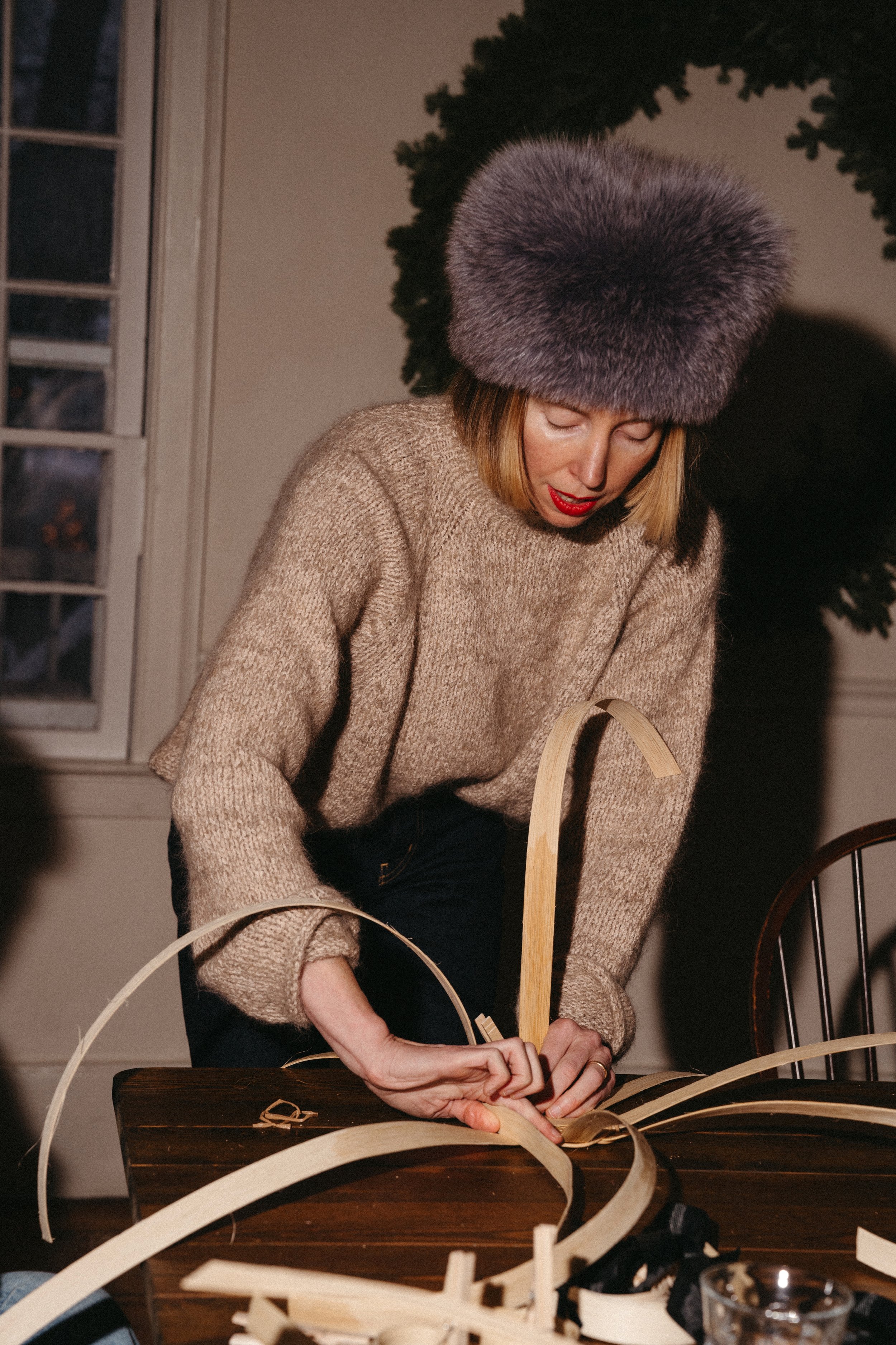 Woman in a beige sweater and gray fur hat shaping a wooden craft at a table.