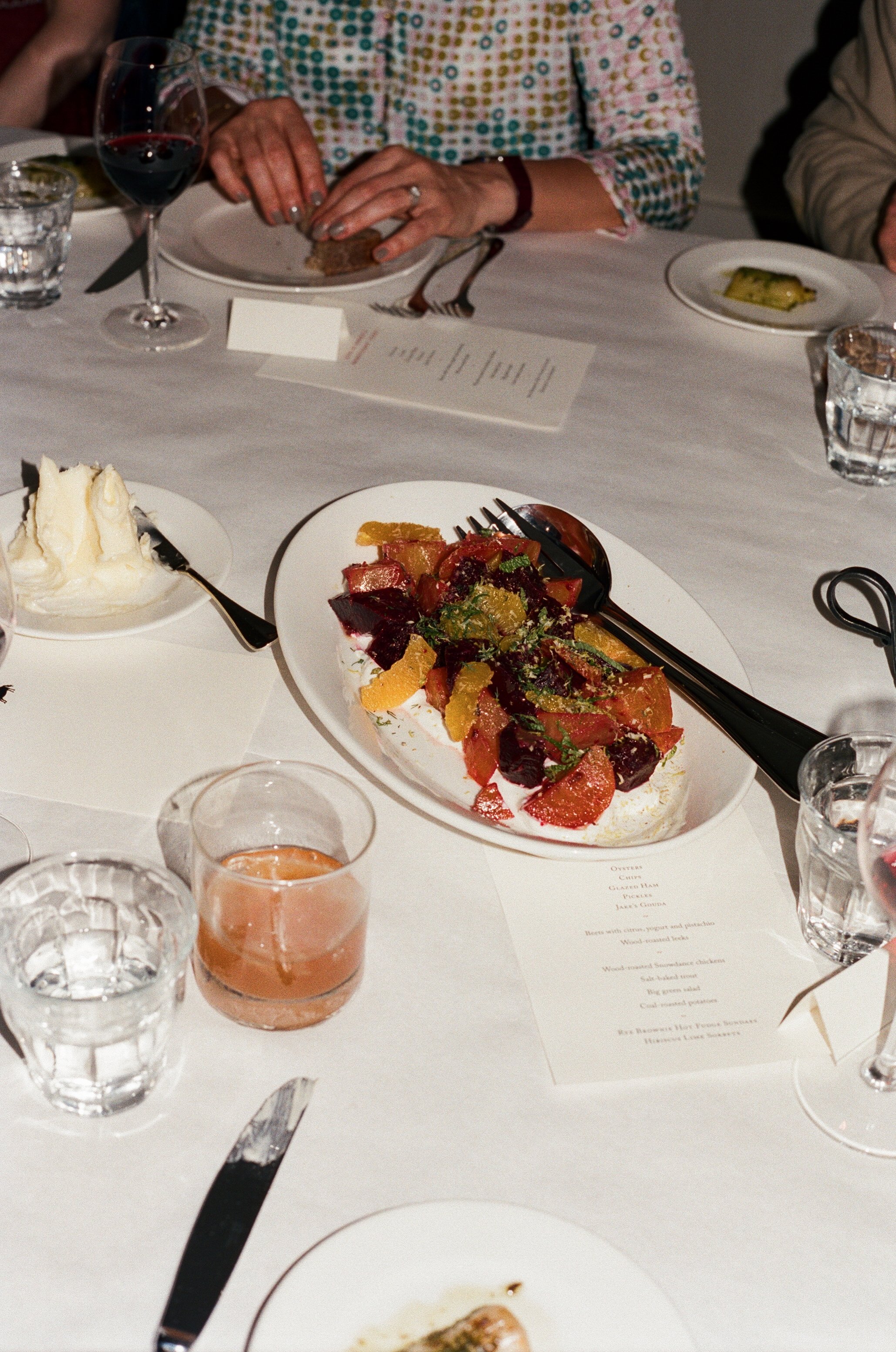 A dinner table with a salad topped with citrus fruits and greens, a glass of red wine, a glass of peach-colored drink, a slice of bread with butter, and multiple glasses of water, set for a meal with several people.