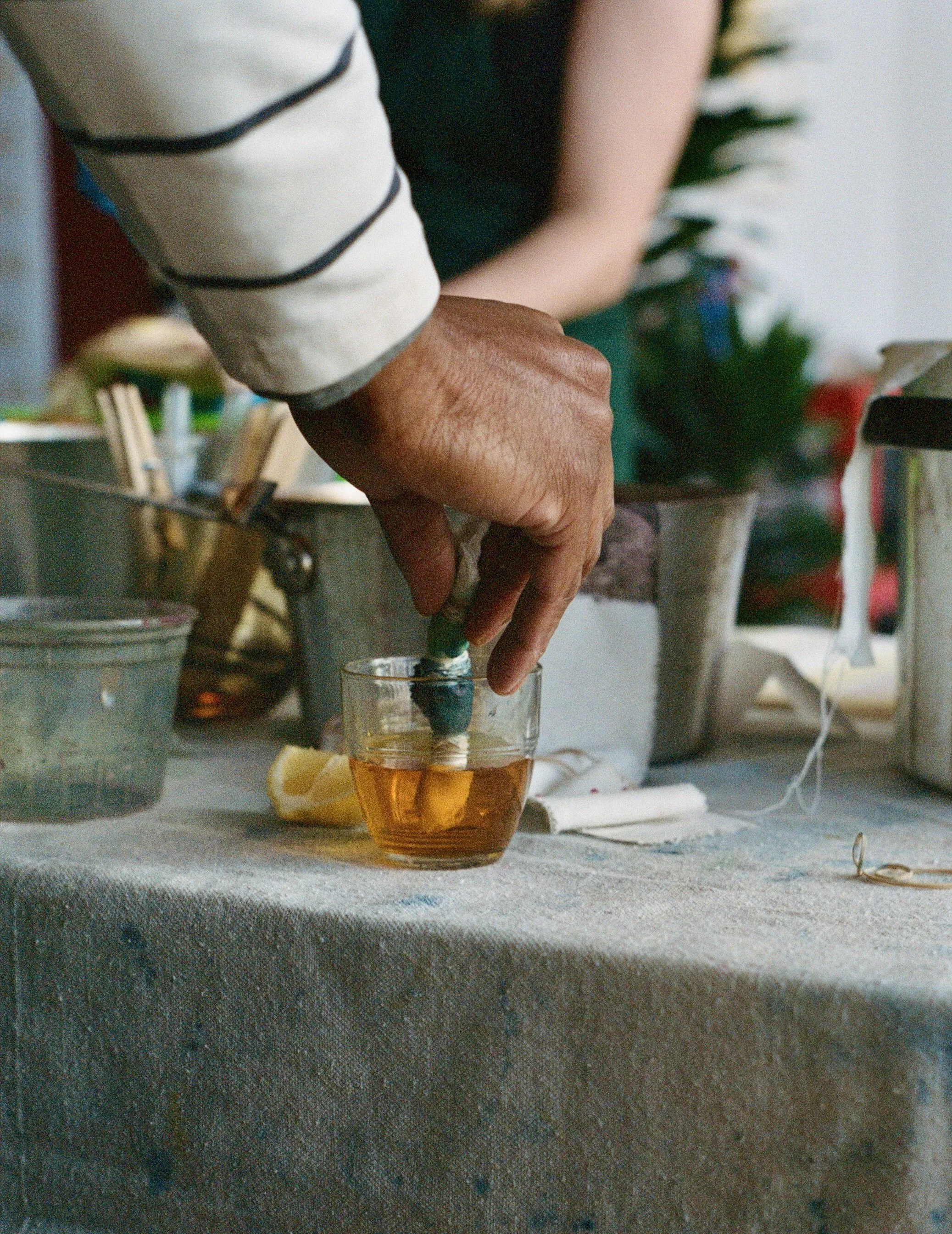 A person's hand drops a green capsule into a glass of amber-colored liquid on a cluttered table.