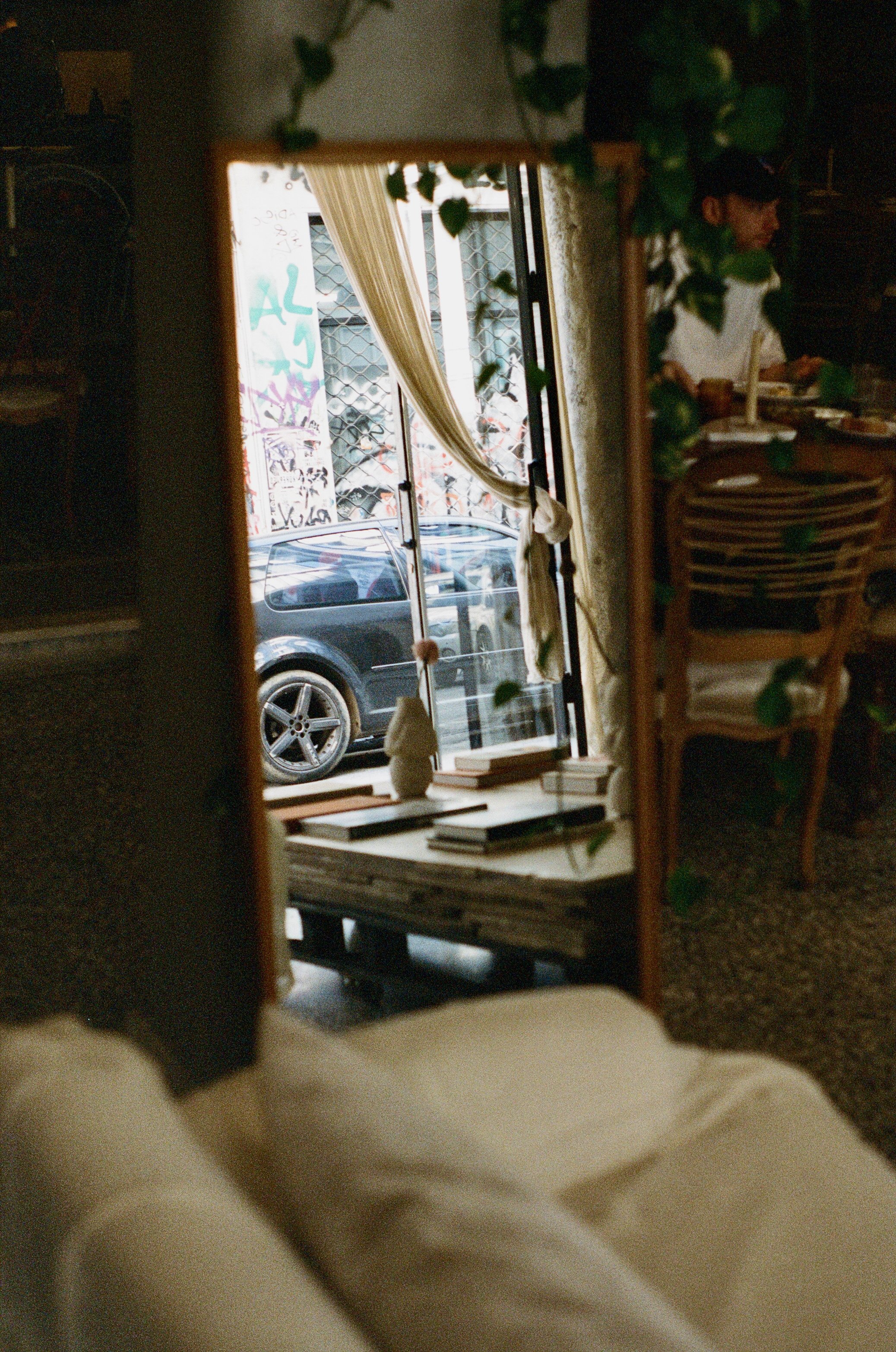 Interior view of a cozy cafe or restaurant, seen through a tall mirror. The mirror reflects a window with cream-colored curtains, a parked black car outside, and graffiti on the outside wall. Inside, there are tables and chairs, one with a person hav