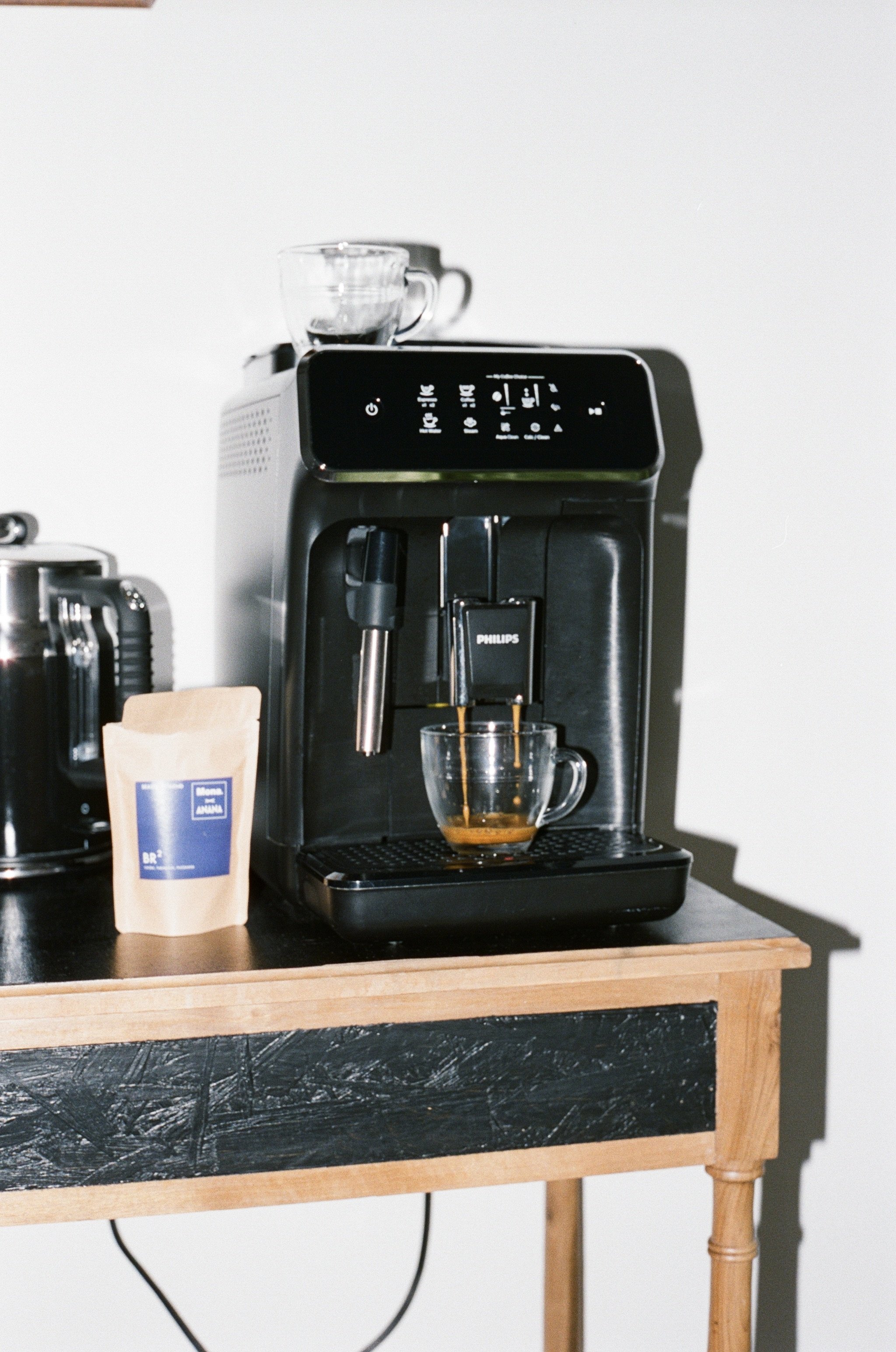 Black coffee espresso machine with a shot pouring into a glass cup, on a wooden table with a packet of coffee beans nearby.