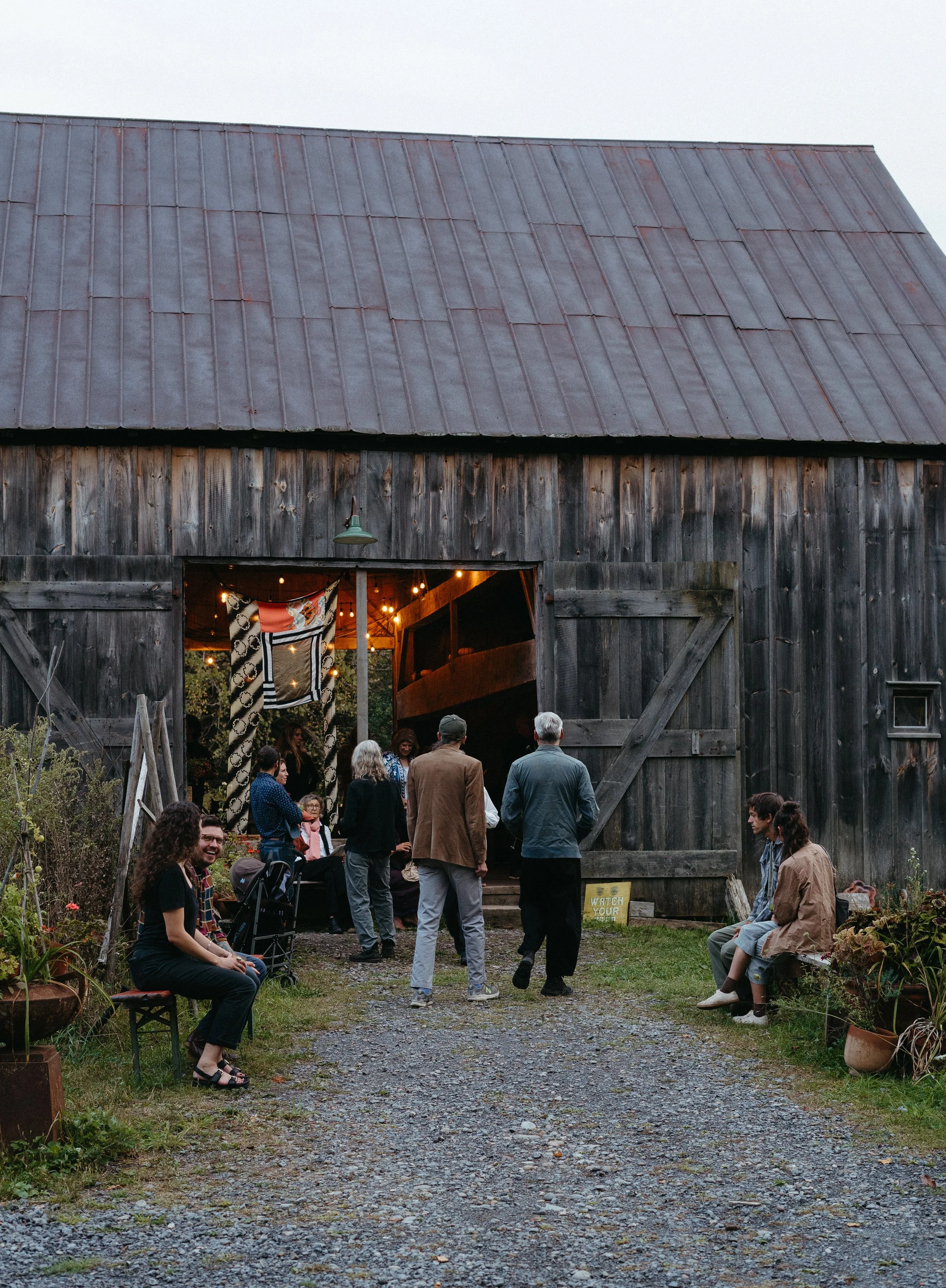 People gathered outside a rustic barn, with some sitting on benches and others standing, as the barn is decorated with string lights and banners inside.