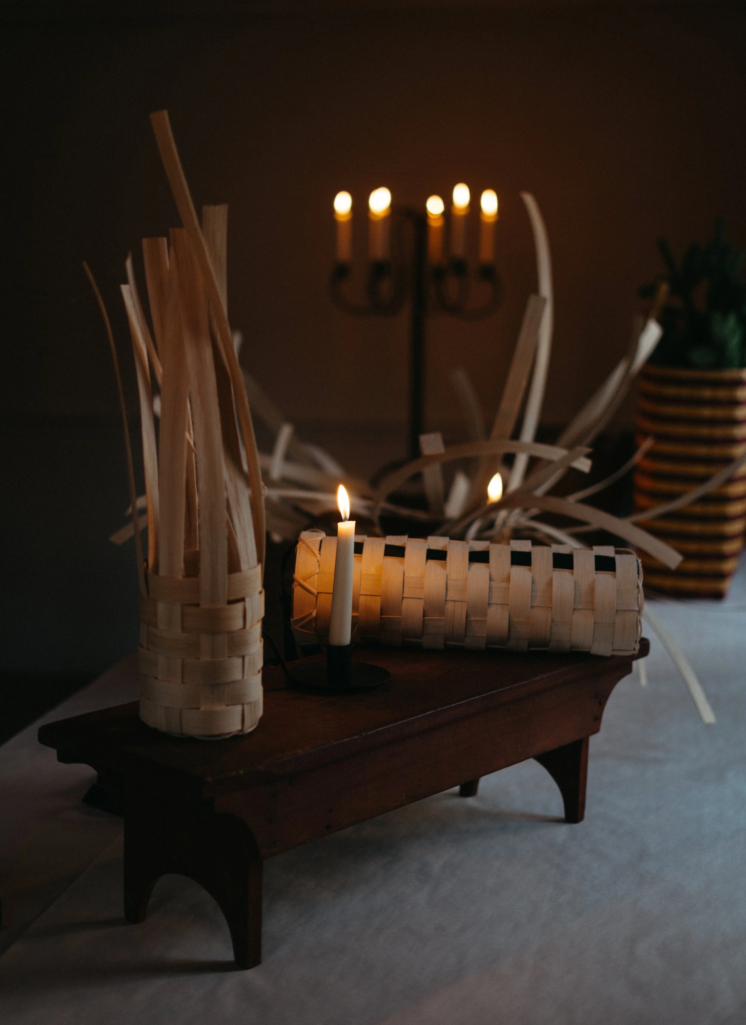 Dimly lit room with a small wooden table, a single candle, woven baskets, a potted plant, and a candelabra in the background.