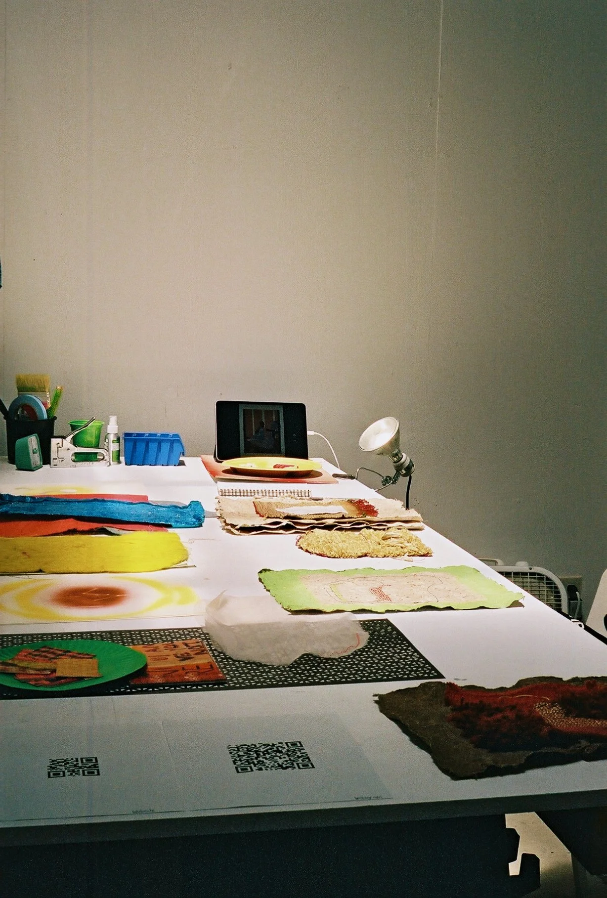 Table with various fabric pieces, tools, a lamp, and a tablet, in a neutral-colored room.