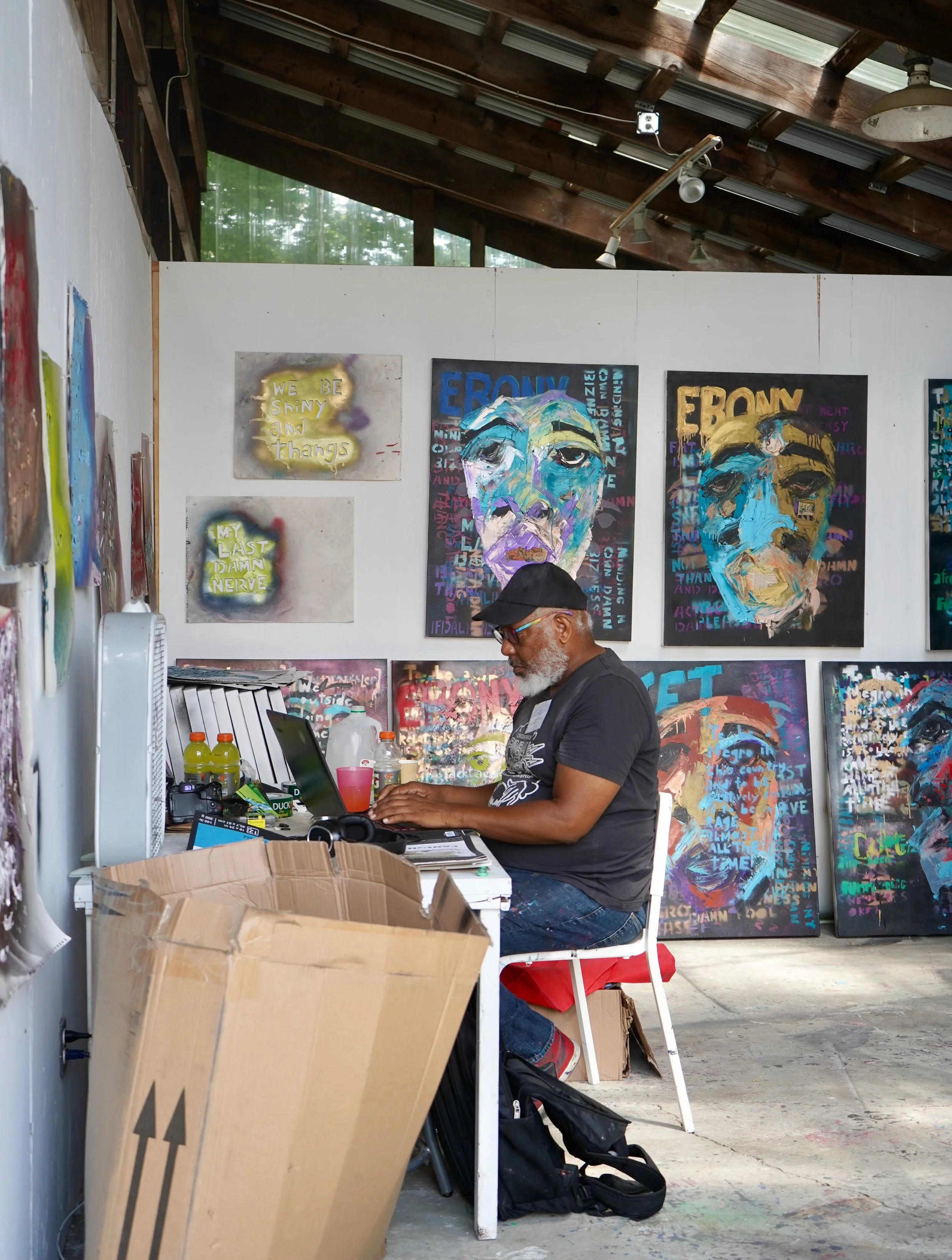 An artist in a black t-shirt, glasses, and a cap sitting at a cluttered table in an art studio, surrounded by colorful portrait paintings and graffiti-style artwork on the walls.