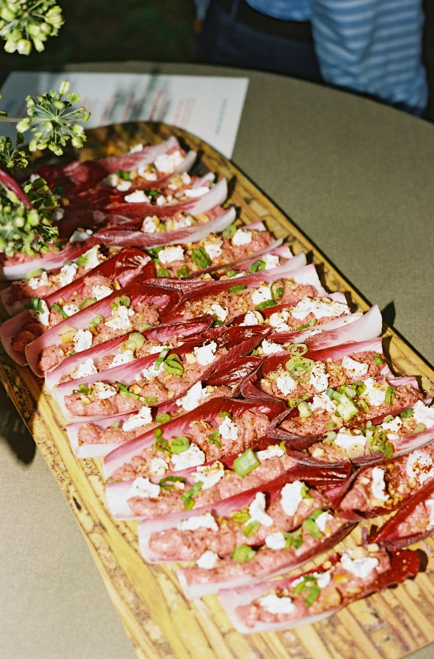 Close-up of a wooden platter with grilled endive topped with a pinkish minced meat mixture, crumbled white cheese, chopped green onions, and small green herbs.