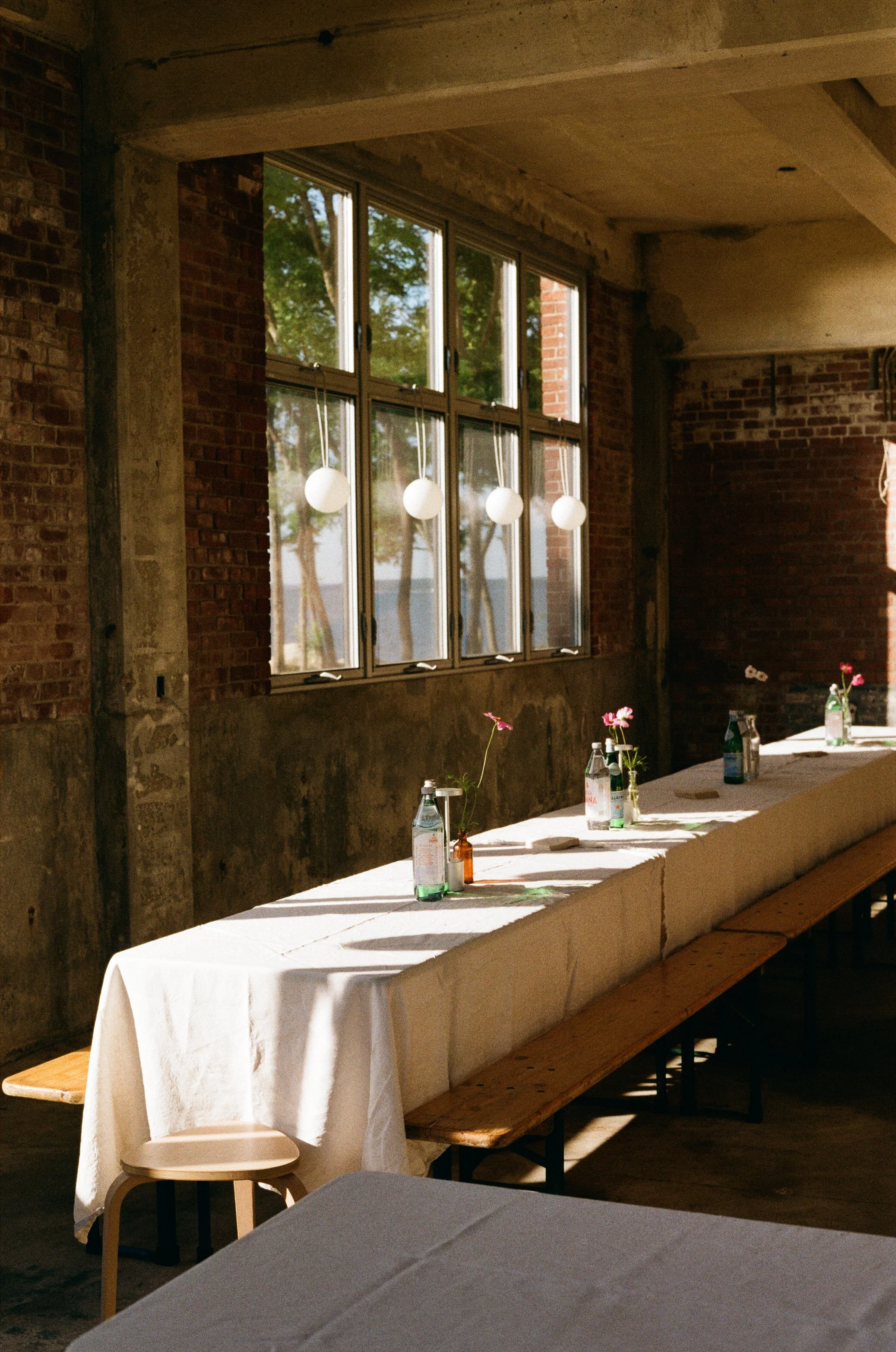 Interior of a rustic event space with a long table covered in a white tablecloth, small bottles with pink flowers, surrounded by benches and a small stool, large windows, and hanging globe lights.