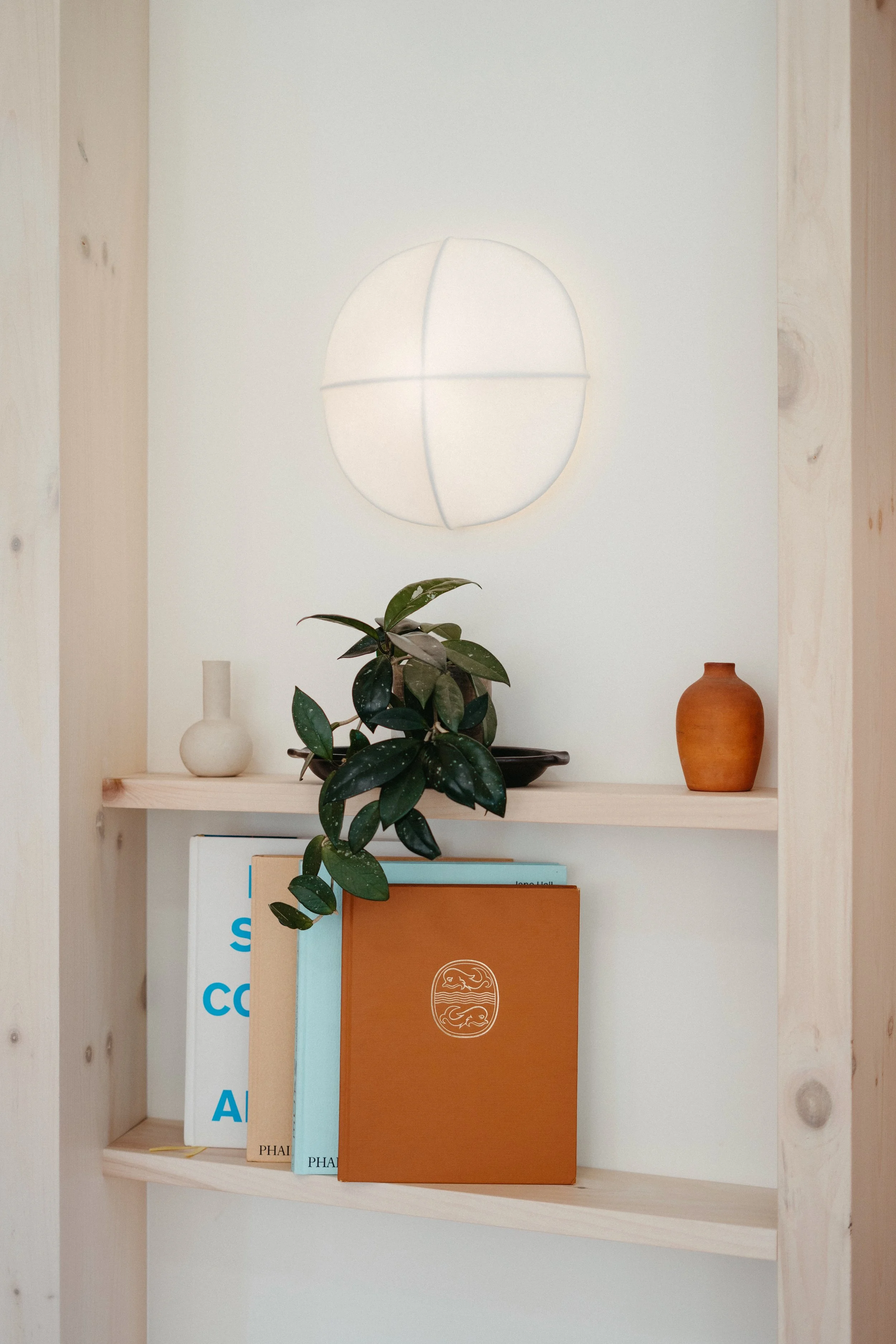 A minimalist bookshelf with books, a green plant, and decorative vases against a white wall with a circular light fixture.