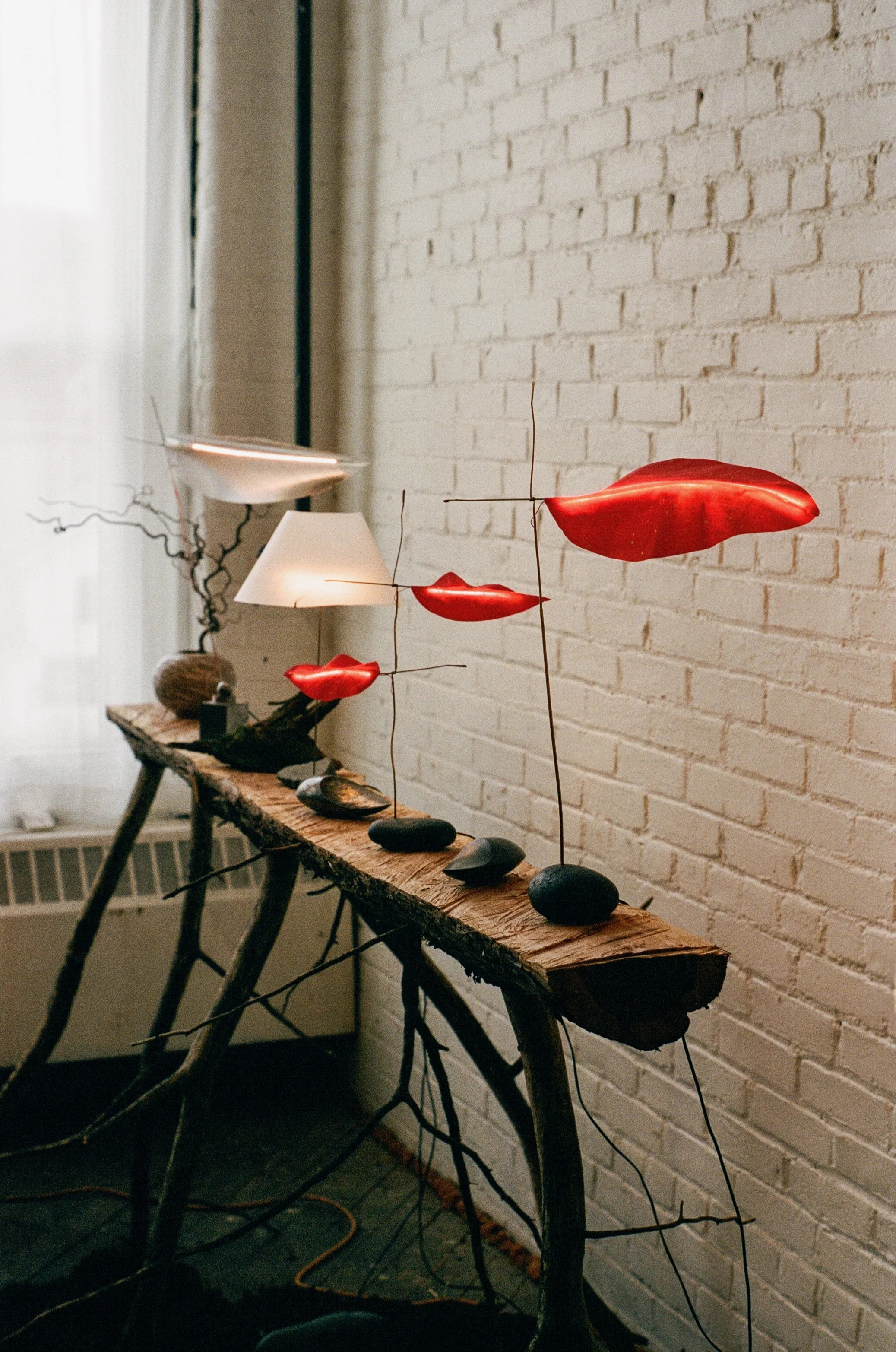 Artistic sculpture with red lips mounted on metal rods, arranged on a wooden table with rocks, set against a white brick wall in a room with a window and curtain.