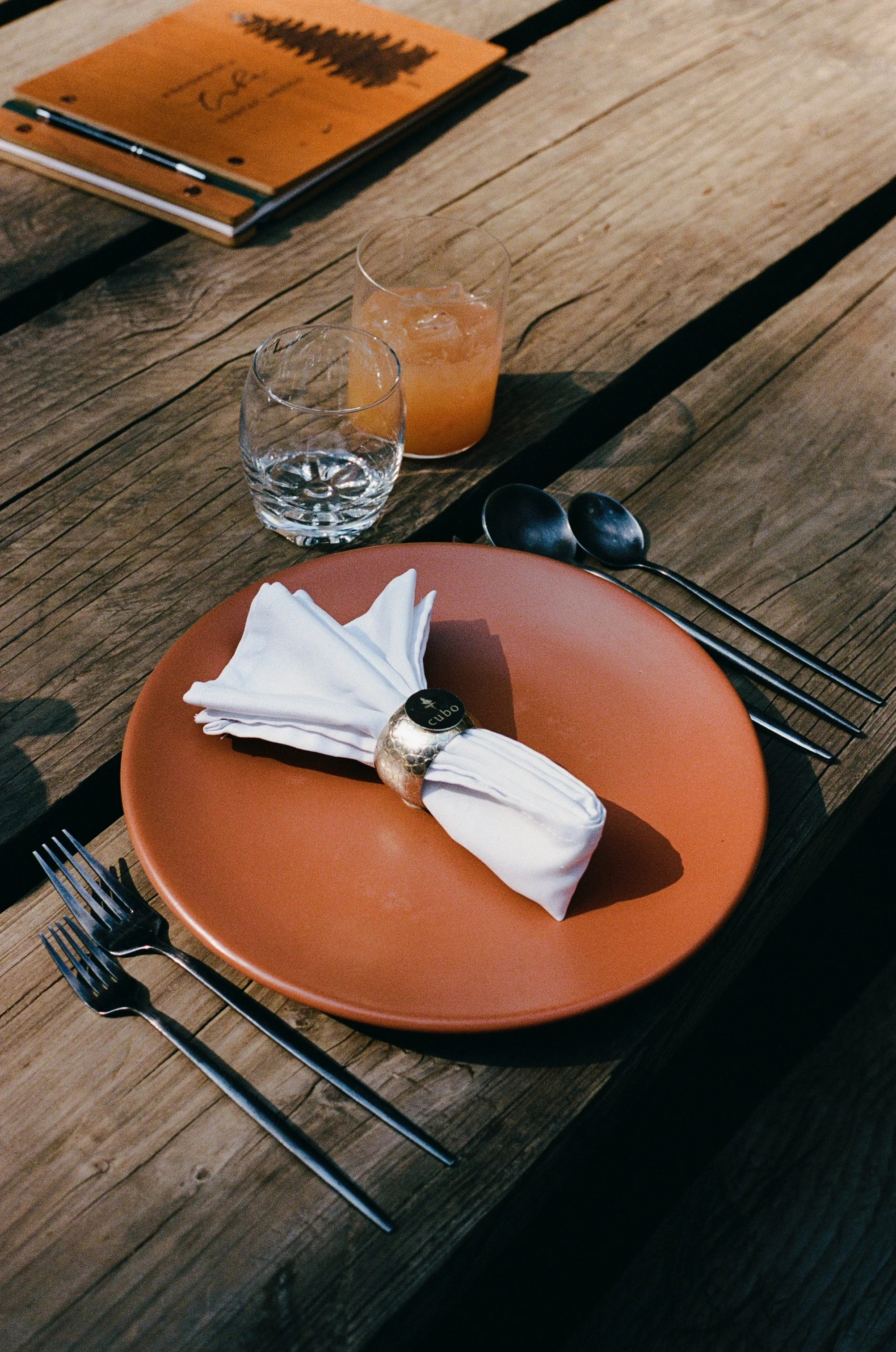 Table setting with a rust-colored plate, a folded white napkin secured with a gold ring, two forks on the left, two black spoons on the right, a glass of water, a glass of orange juice with ice, and a menu on a rustic wooden table.