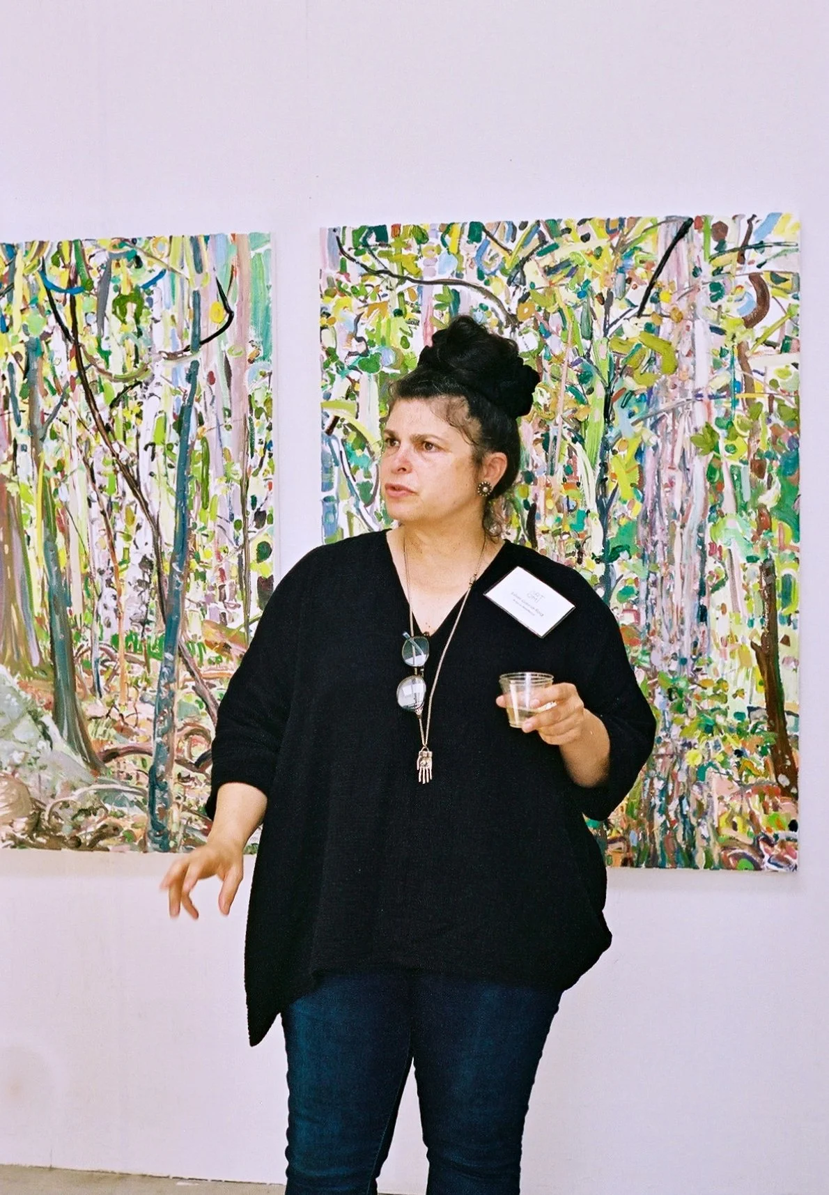 A woman at an art gallery standing in front of colorful abstract paintings, holding a small cup, wearing a black sweater, a name badge, and a long necklace.