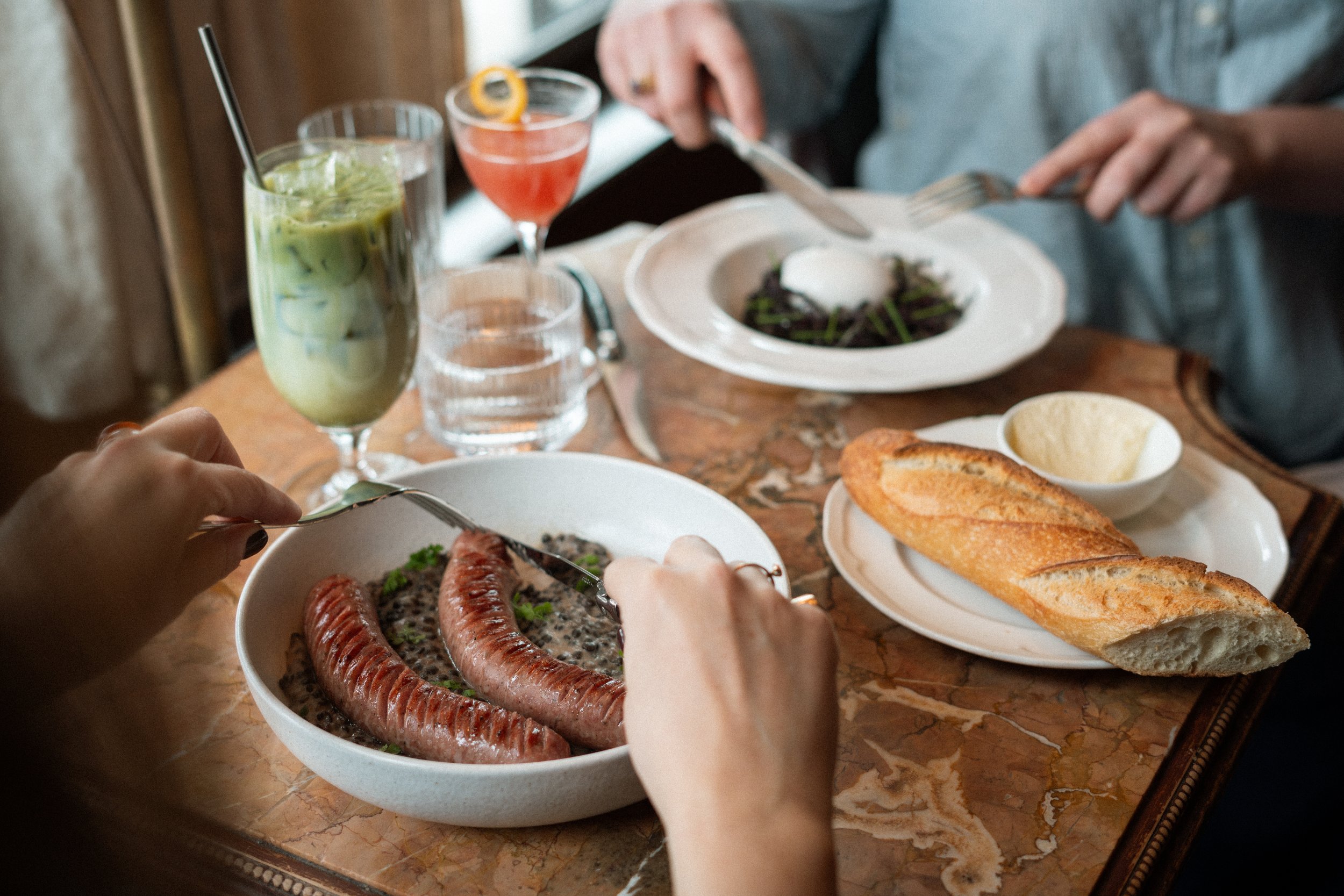 People dining at a table with sausages in a bowl, baguette bread, salads, and colorful drinks.