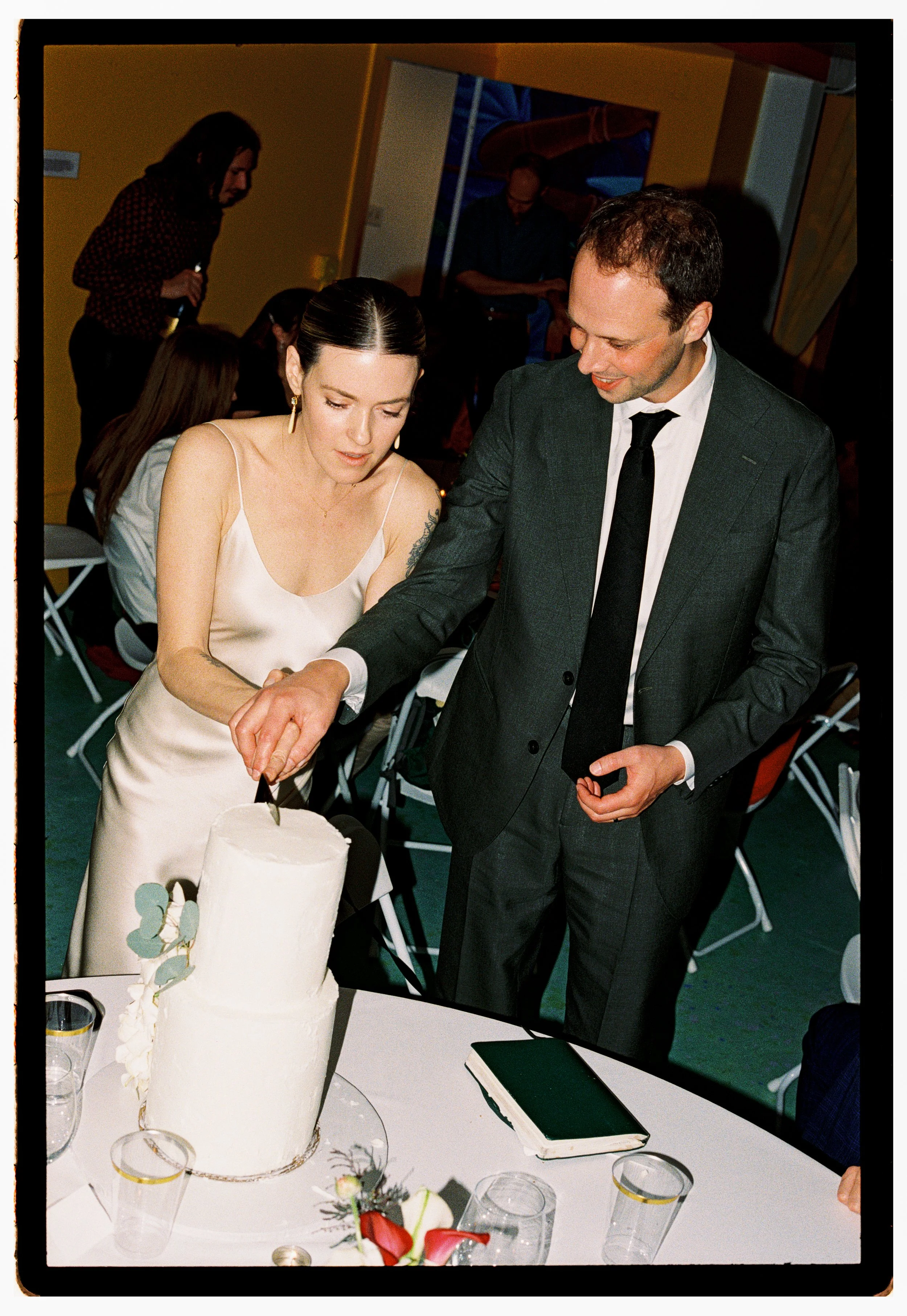 A woman in a white dress and a man in a suit are cutting a white wedding cake together at a celebration. There are glasses and flowers on the table in front of them, and other people are visible in the background.