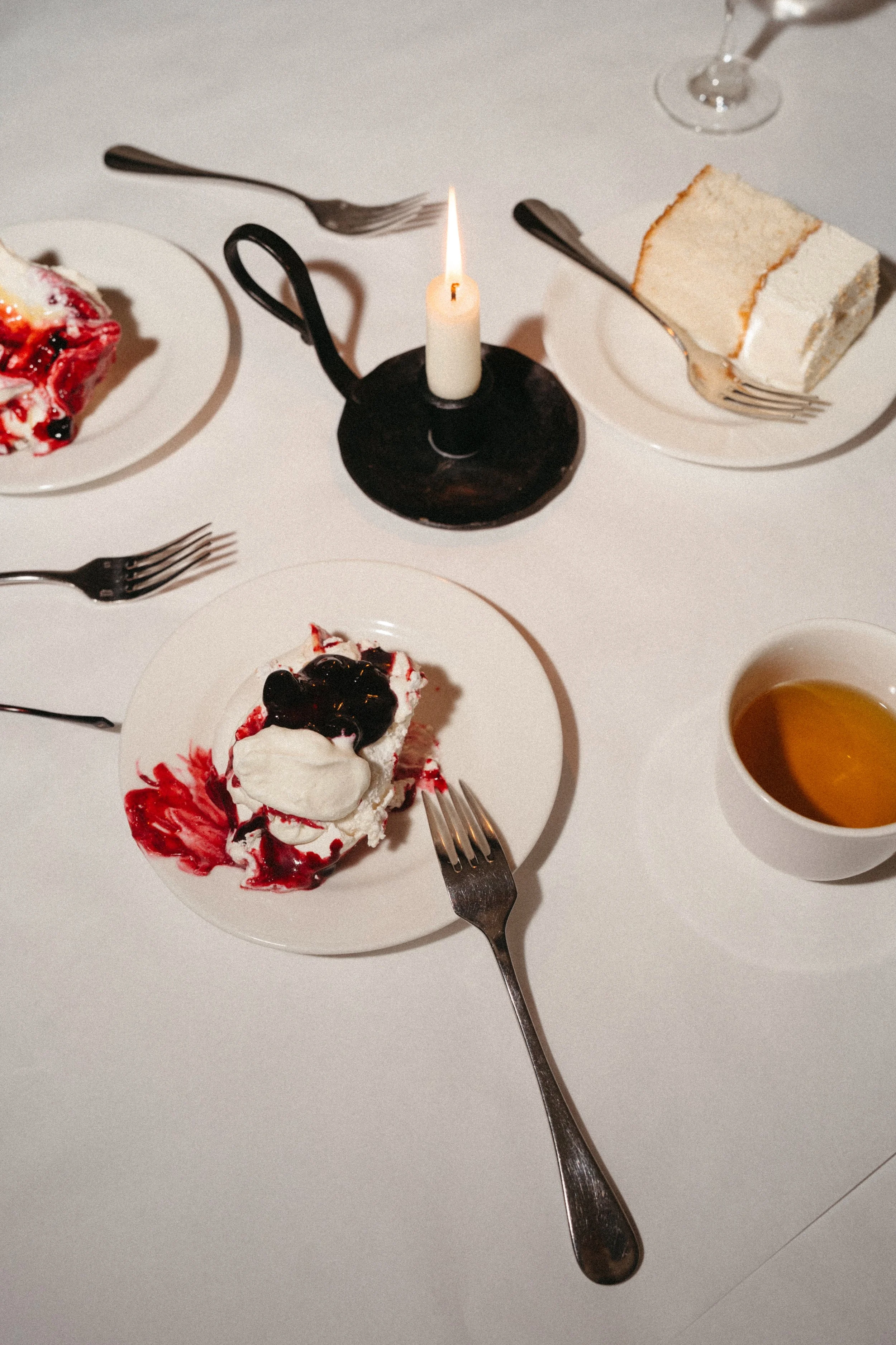 A white table setting with a slice of cake, a dessert with berries and whipped cream, a cup of tea, and a candle in a black candlestick holder.