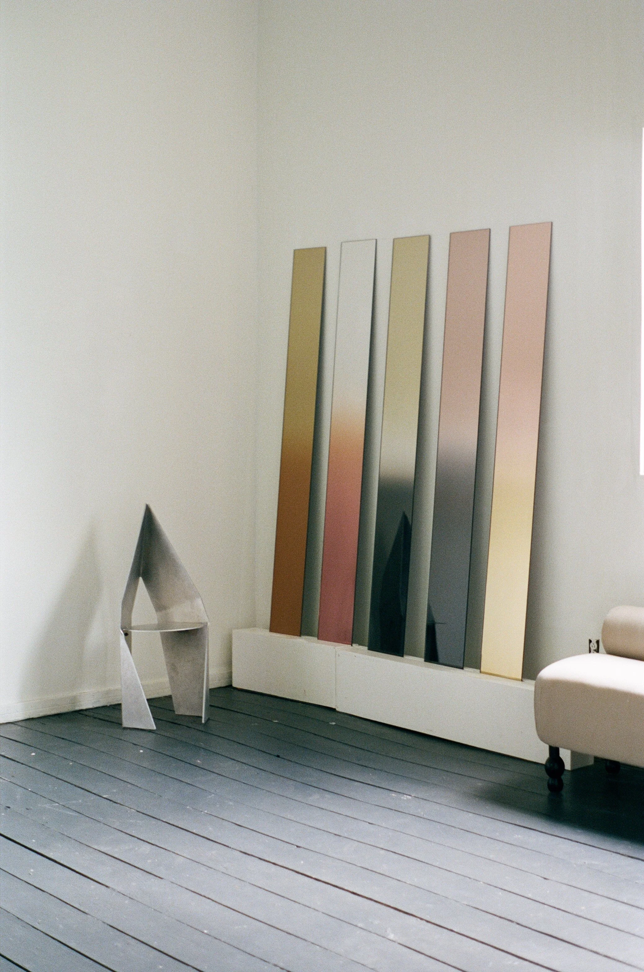 An art gallery corner displaying five tall, narrow abstract mirrors or panels in shades of metallic, pink, green, black, and yellow leaning against a white wall. A modern, angular metallic chair is on the left, and part of a beige sofa is on the righ