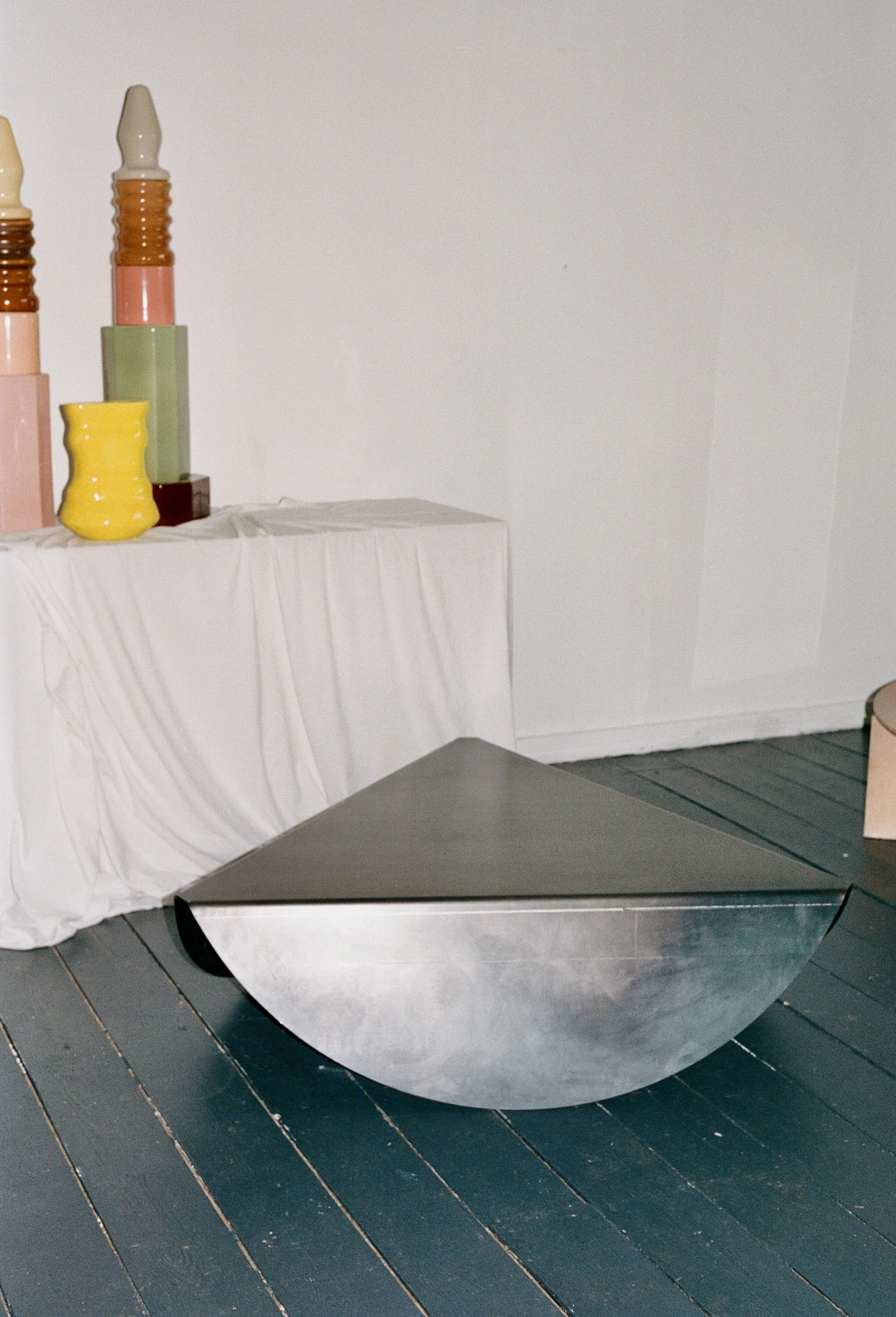 A minimalist art installation featuring a geometric metallic sculpture with a triangular top and half-sphere base, placed on a wooden floor. To the left, a table draped with white fabric displays three large ceramic vases resembling escalator steps i