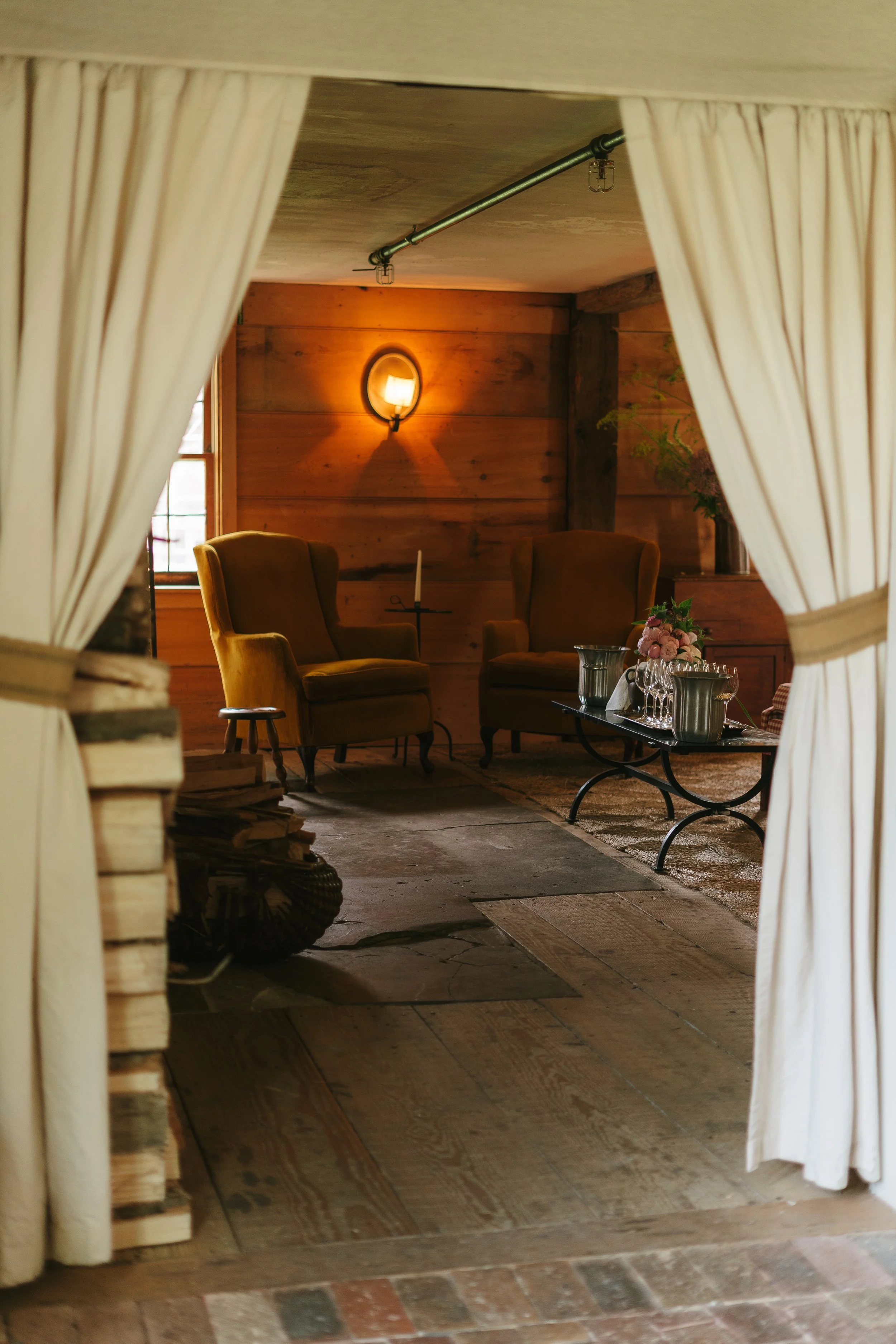 View through a curtain into a cozy rustic seating area with two armchairs, a side table, and a coffee table with flowers and glassware, illuminated by warm wall lighting.