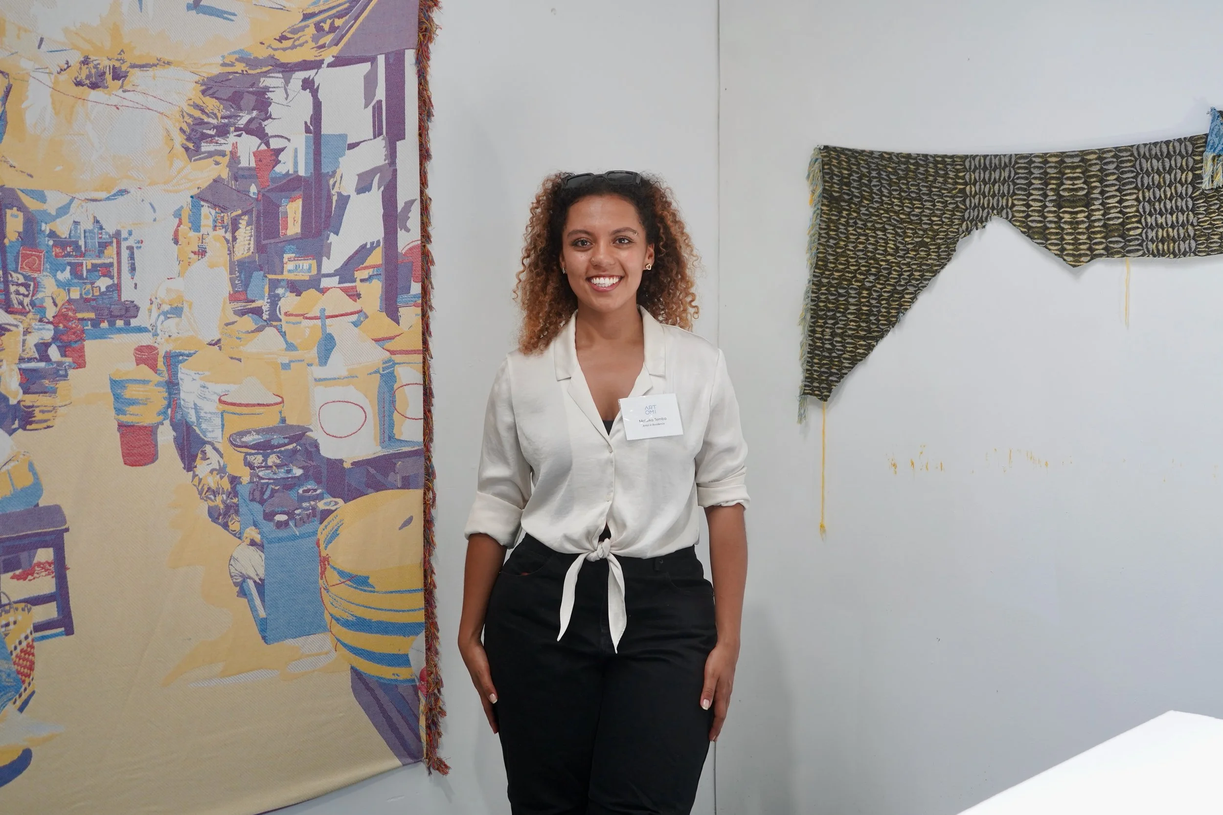 A woman standing in an art gallery, smiling, with artwork hanging on white walls behind her.